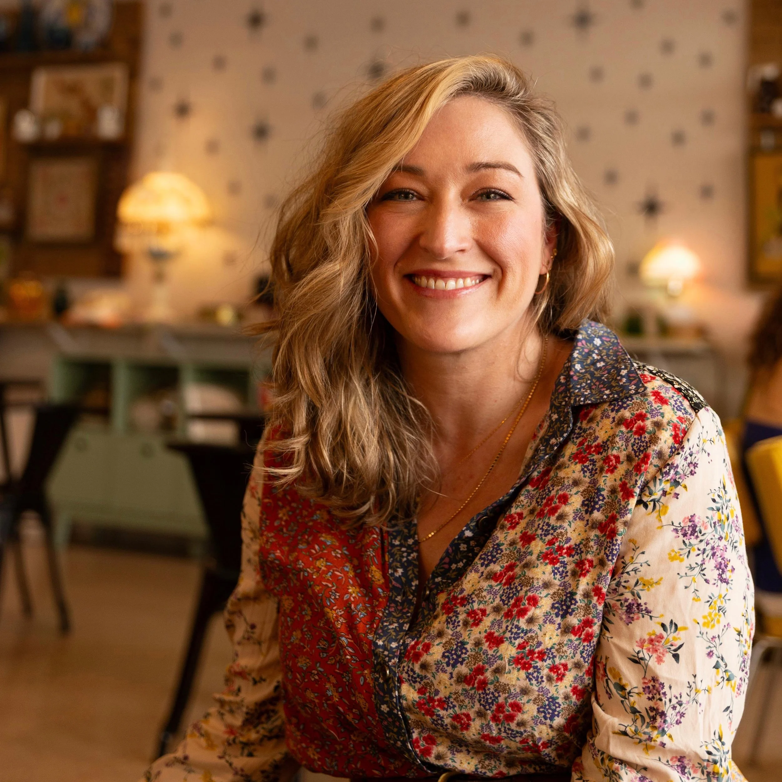 A woman with long, wavy blonde hair and light skin smiling, sitting on a yellow couch with a black cardigan and a floral dress, posed in front of a white wall and ornate framed mirror.