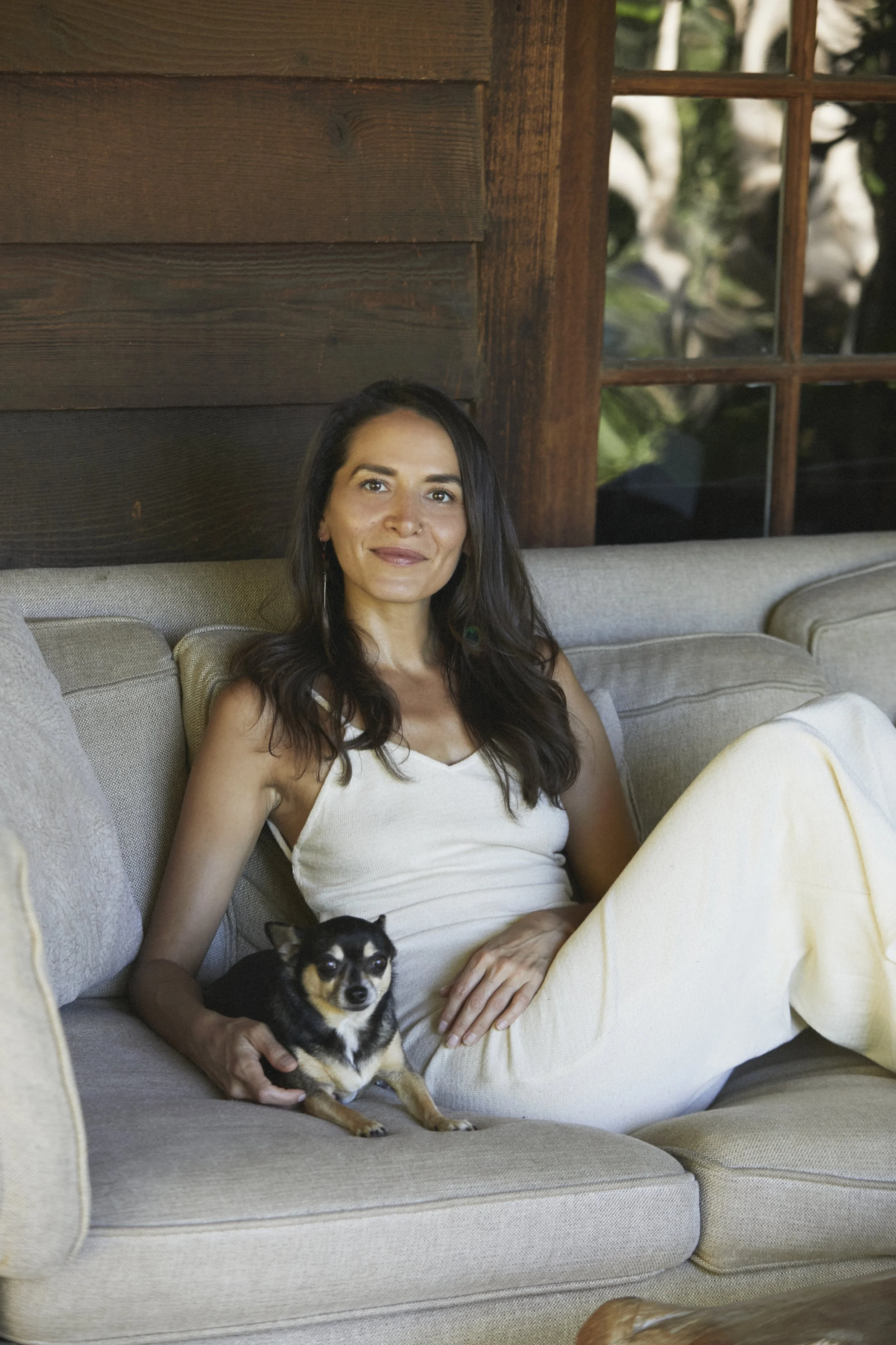 A woman with long dark hair sitting on a beige sofa with white cushions, holding a small black and tan dog, in a room with wooden walls and a window showing greenery outside.