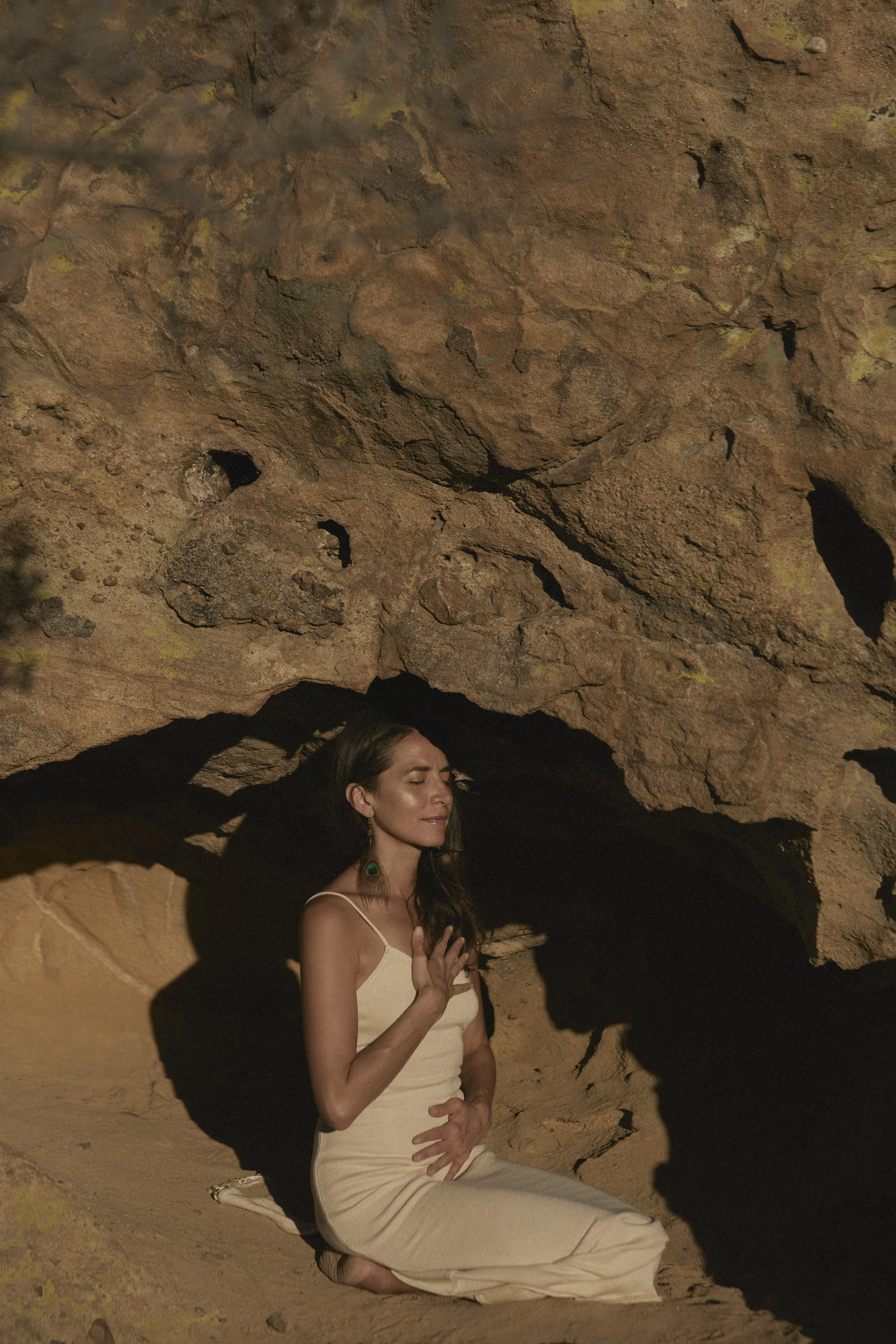 A woman wearing a cream-colored dress is kneeling on the ground with her eyes closed, in a cave or rocky area with a prominent rock formation overhead creating a shadow.