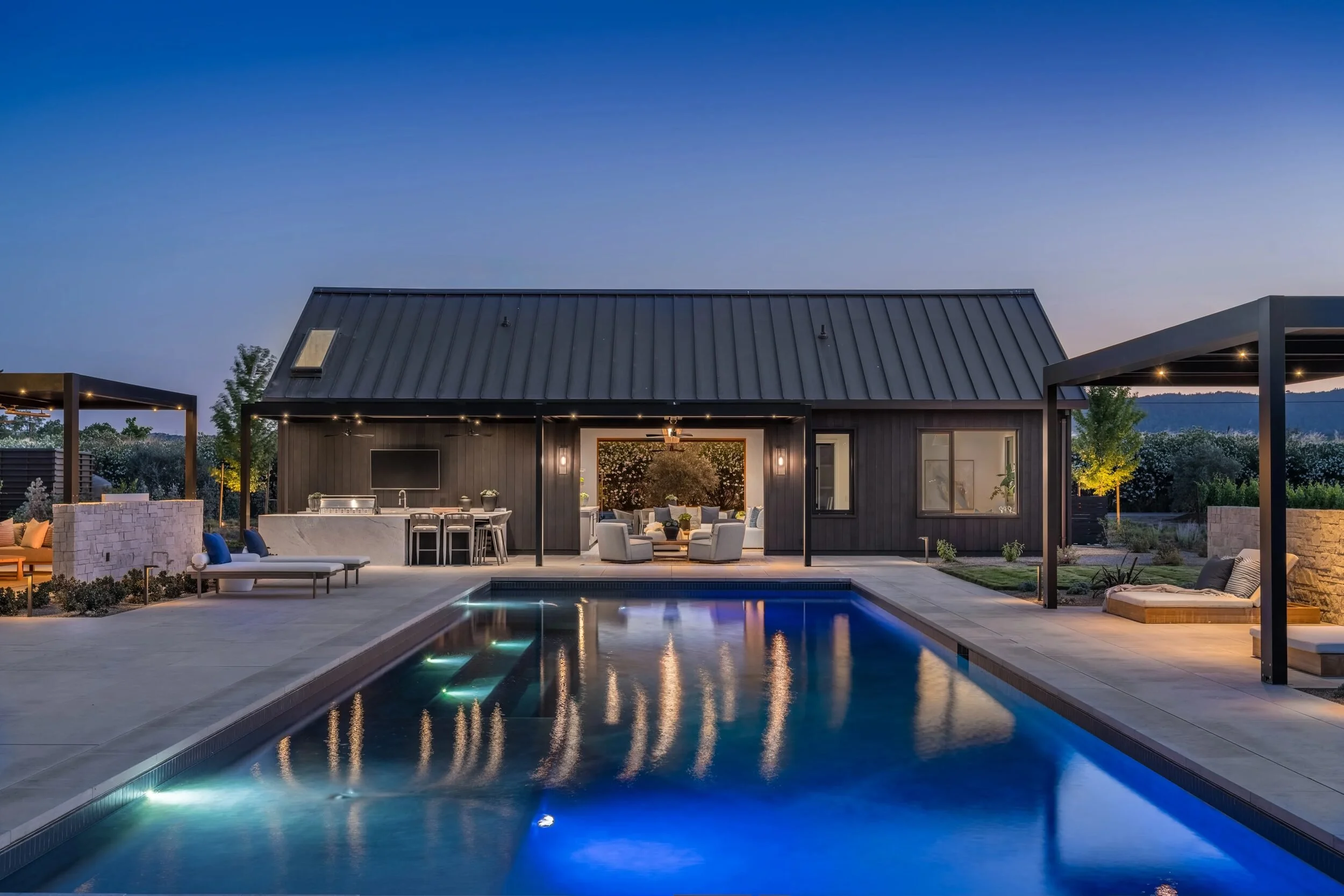 pool house at dusk with outdoor kitchen, covered patio, pergolas, and illuminated pool