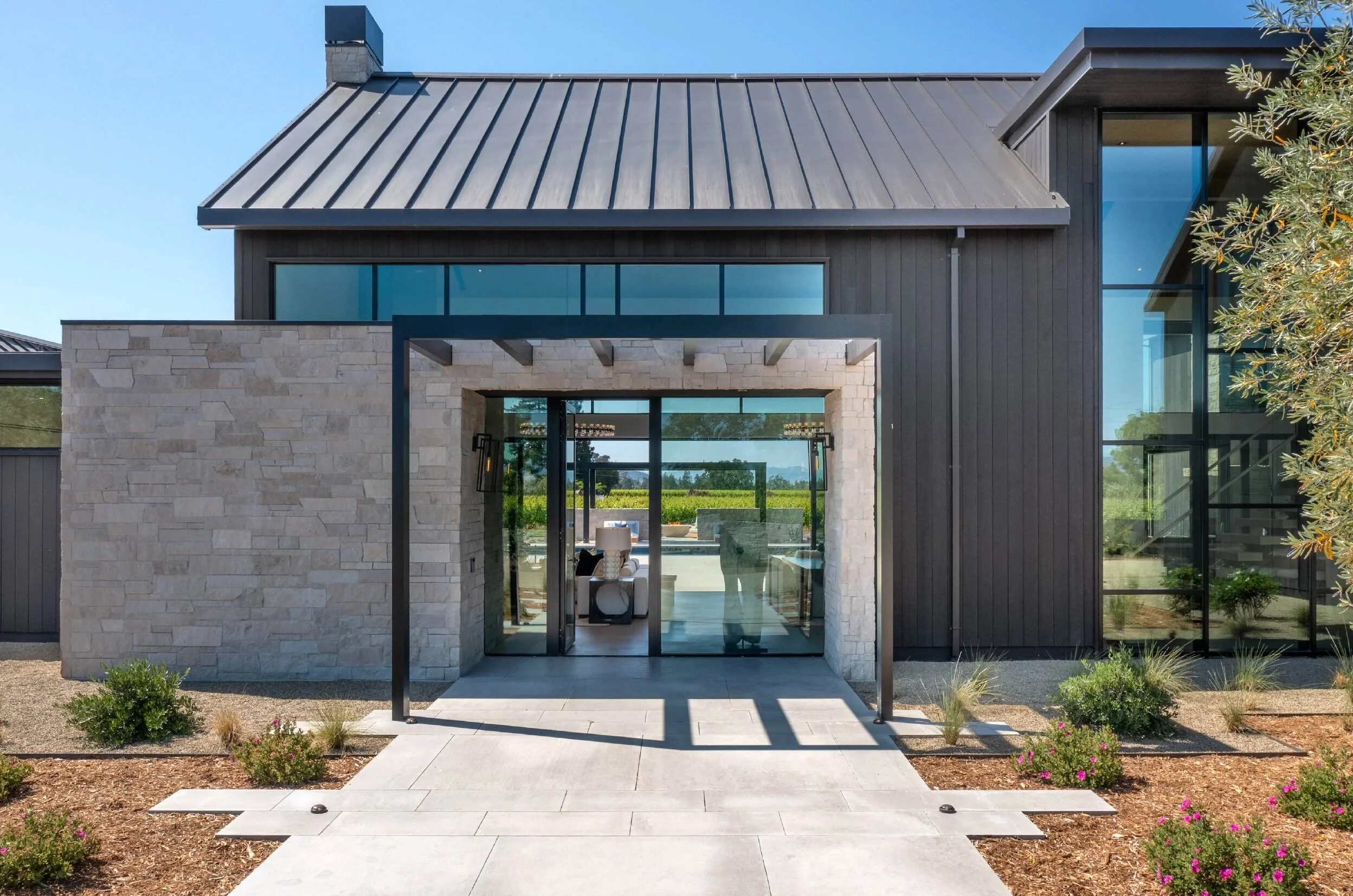 Stone-clad entry with steel canopy and glass doors framing vineyard view beyond