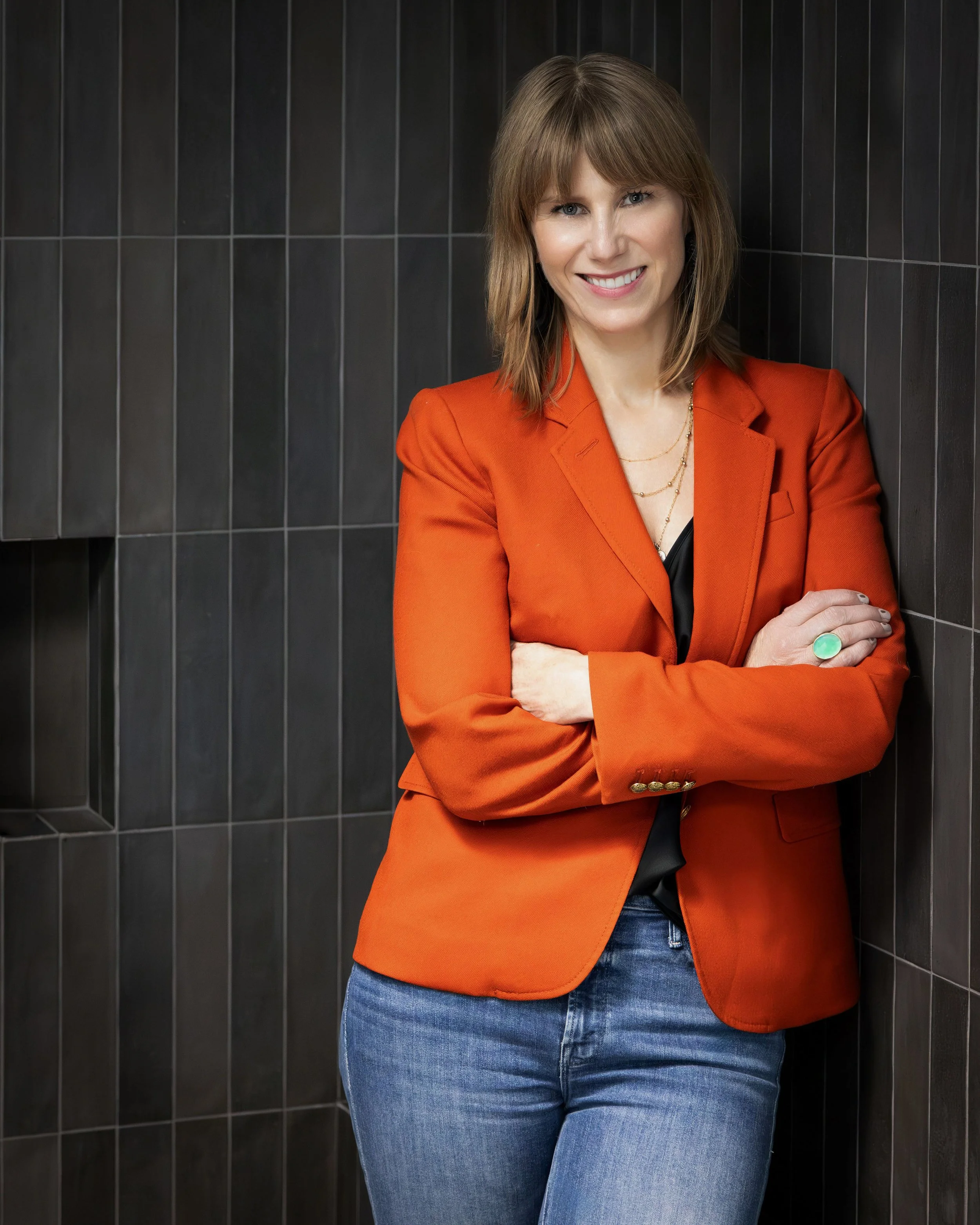 Image of Anne of Anne leaning against charcoal tile wall wearing bright orange jacket.