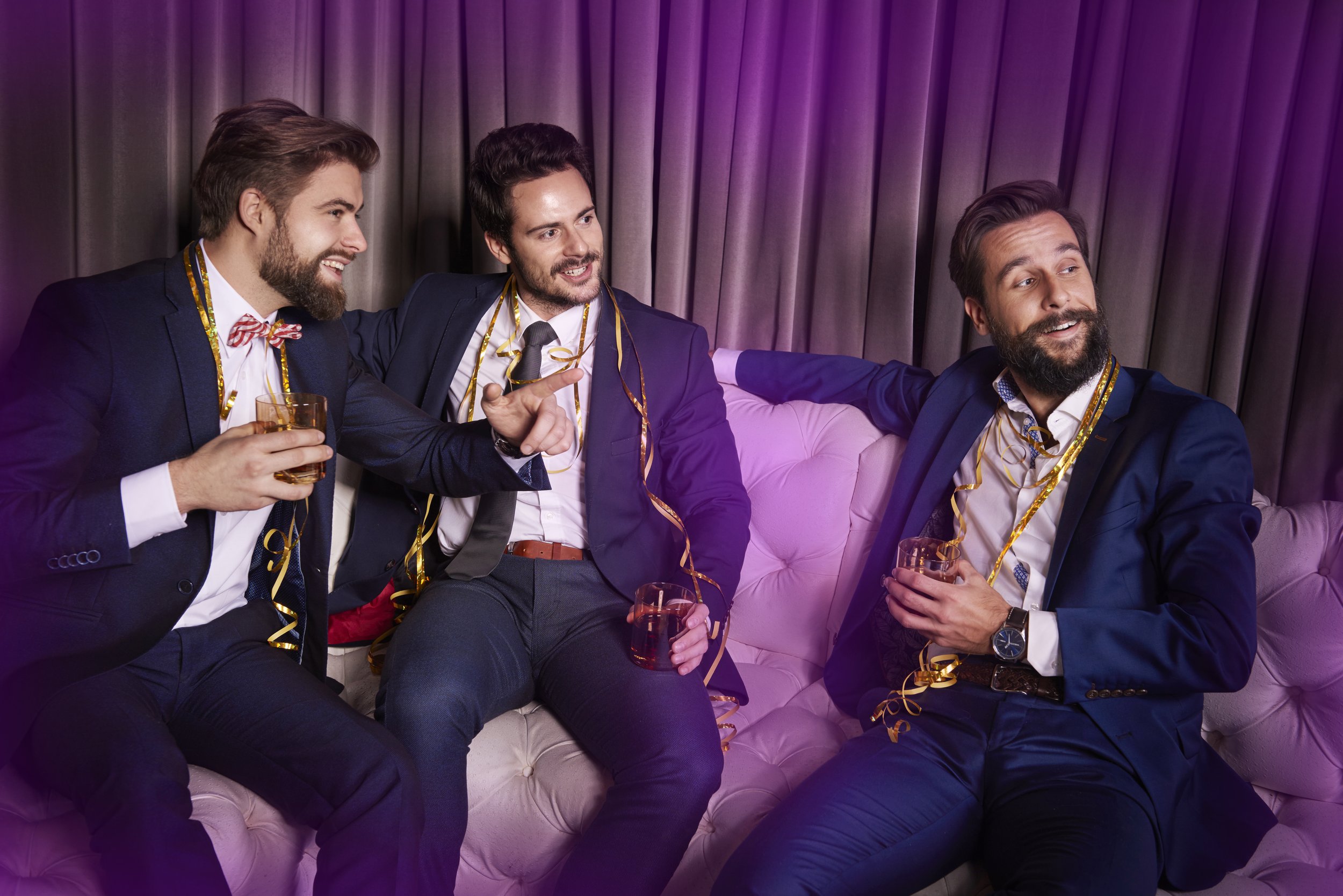 Three men gather on a pink sofa, wearing suits, with party hats and decorations, holding drinks, celebrating at a party with purple curtains in the background.