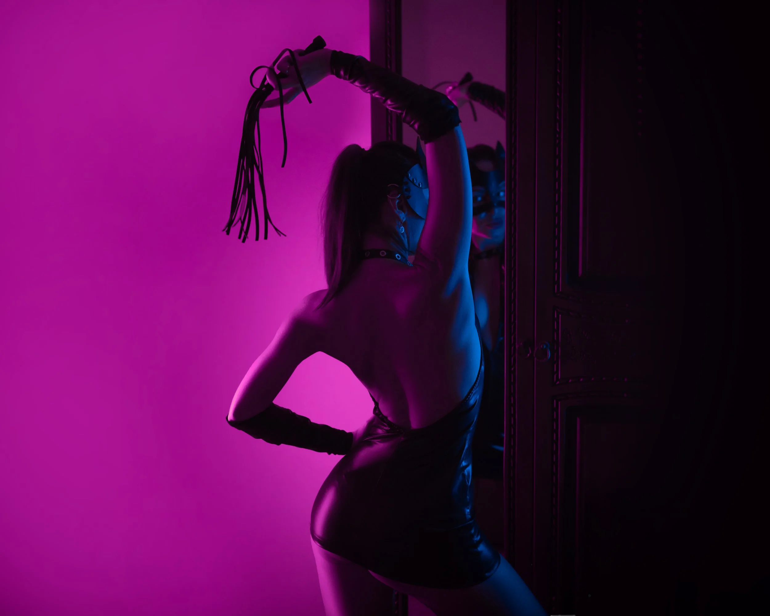 A person with long hair in a ponytail wearing black gloves, a face mask, and leather pants, holding a whip, standing in front of a mirror with purple and pink lighting.