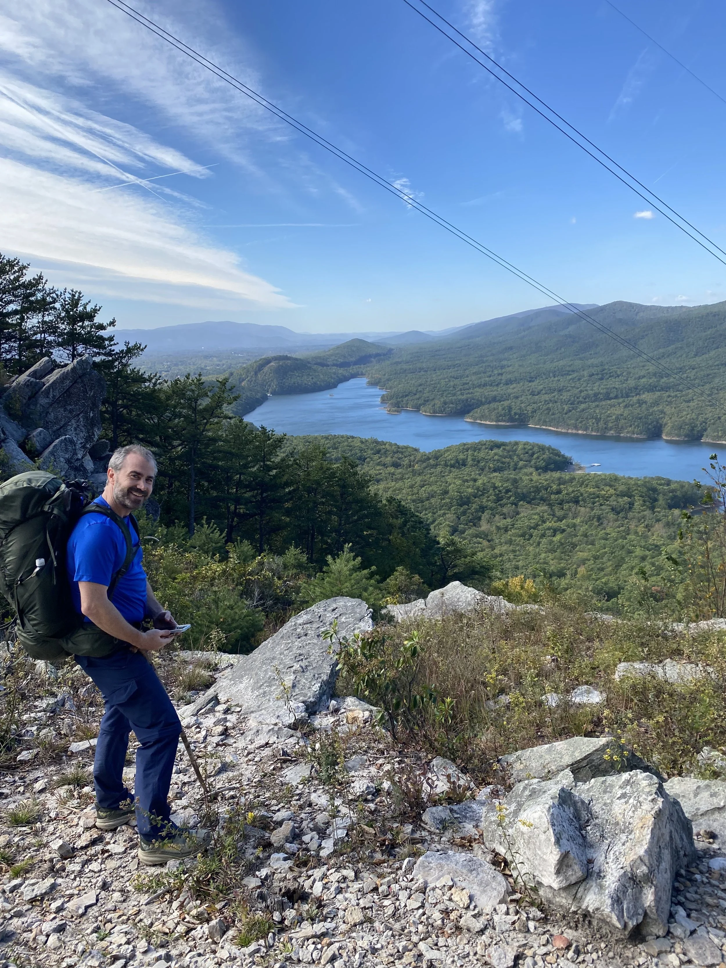 A hiker standing on rocky terrain with a backpack, overlooking a scenic view of a river winding through green forested mountains under a blue sky with some clouds.