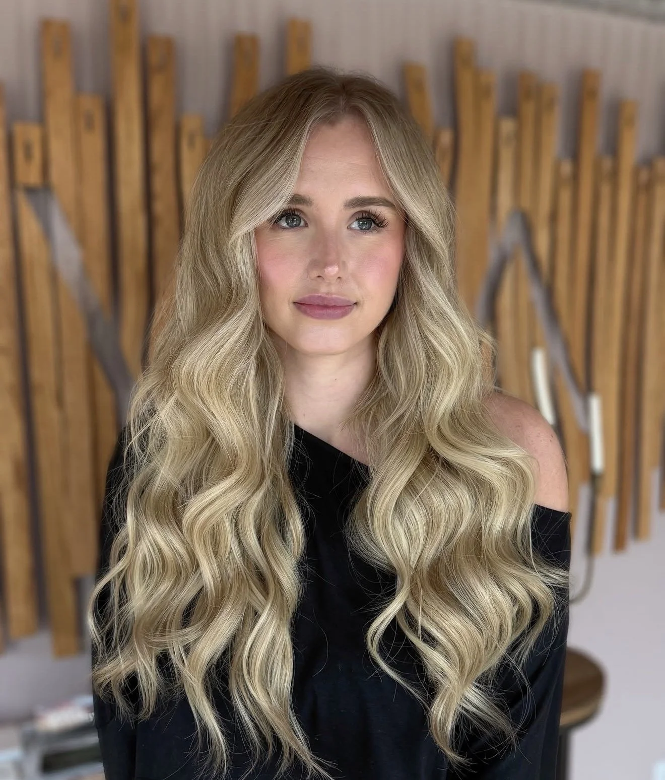 A woman with long, wavy blonde hair and blue eyes, wearing a black off-the-shoulder top, standing in front of a wooden art piece on the wall.