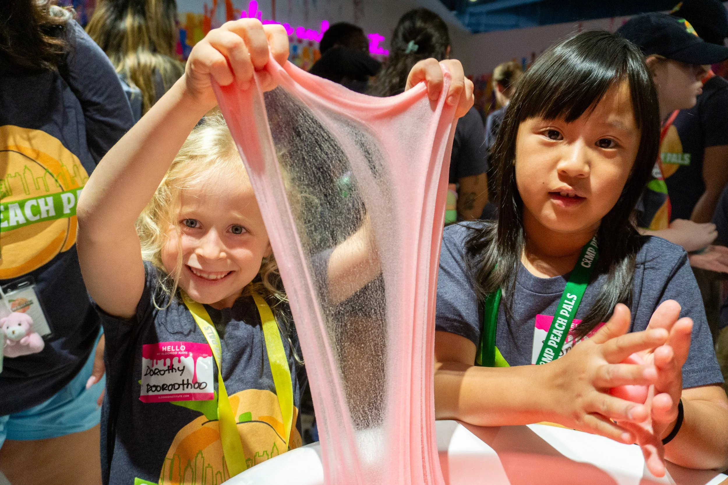 Two young girls, one blonde and one Asian, demonstrate slime on a table. They are part of a group at an event with many people around. The blonde girl is smiling and holding a stretch of pink slime, while the Asian girl watches.