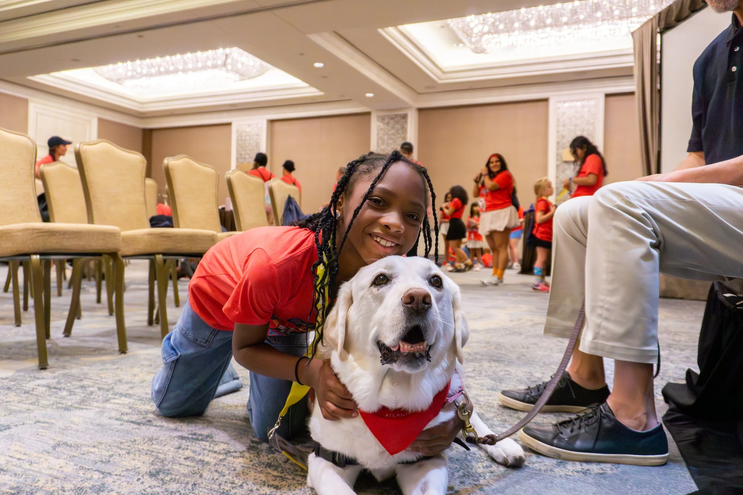 A young girl kneeling and smiling with her arm around a white service dog in a large indoor event space with chairs and people in the background.
