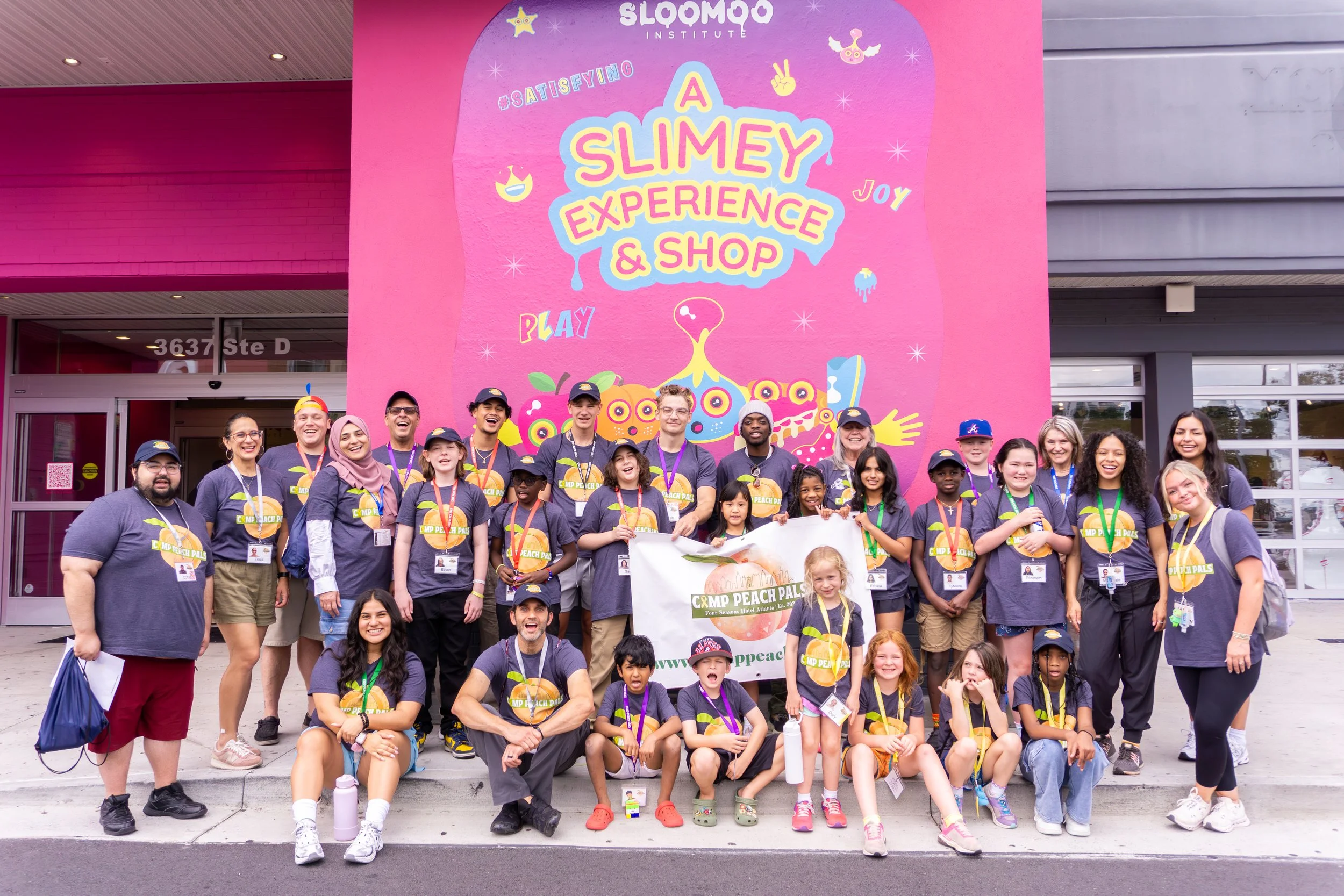 Group of children and adults posing in front of a vibrant pink wall with animated graphics and the words '& A Slimey Experience & Shop'. Many are wearing navy T-shirts with "MP Peach Pal" logos and lanyards, holding a large banner that reads 'CMP PEACH PALS'. The scene appears cheerful and fun, with a diverse group.