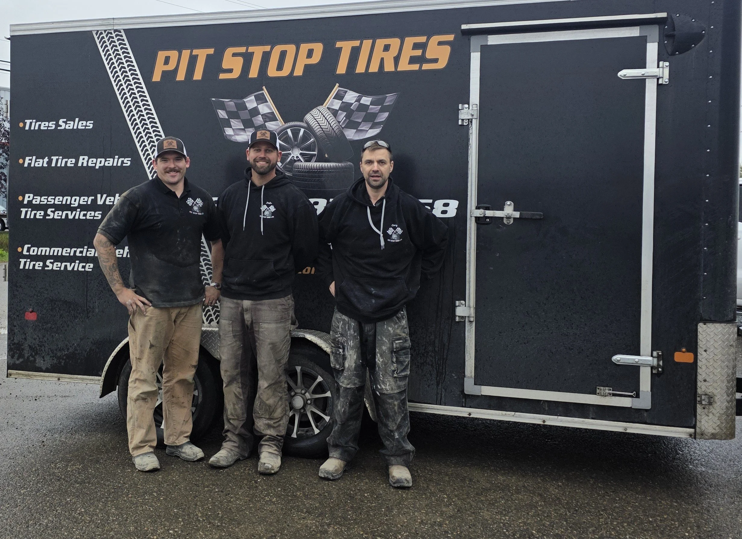 Three men stand in front of a black trailer with the sign 'PIT STOP TIRES' on it. The man on the left, with a beard and mustache, wears a black shirt, tan pants, and a black cap with a logo. The man in the middle, with a beard, wears a black hoodie, beige pants, and a black cap. The man on the right, with short hair and facial hair, wears a black hoodie and camouflage pants. The trailer has graphics of tires and checkered flags, listing services like tire sales, flat tire repairs, passenger vehicle services, and commercial truck services.