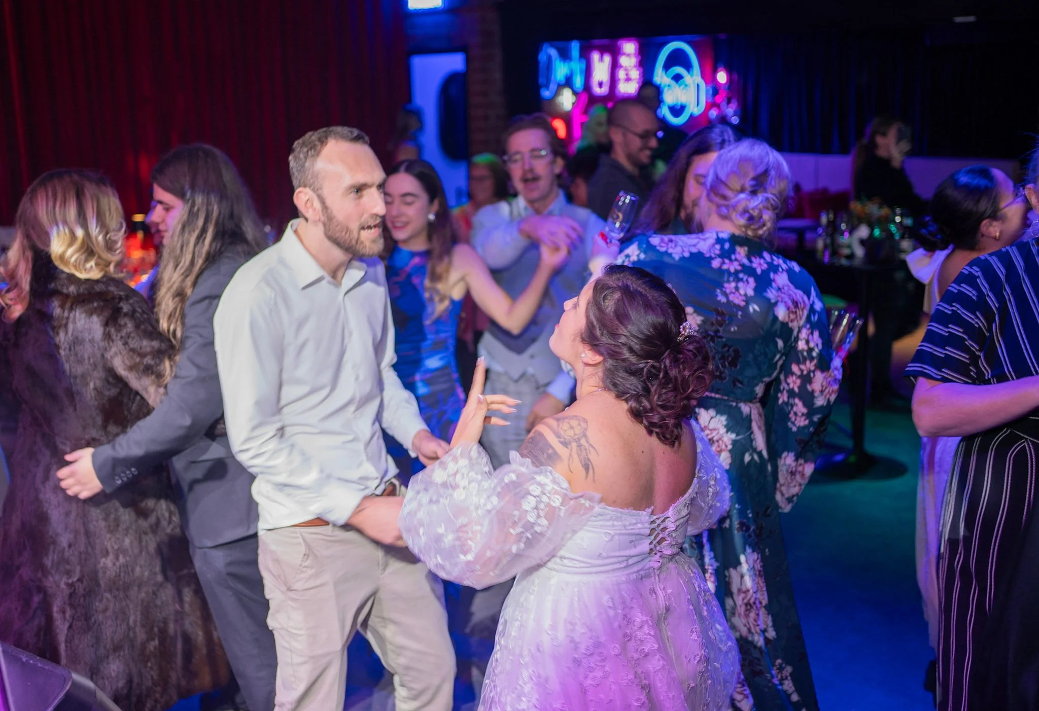 Bride and groom dancing closely with friends in a lively, neon-lit atmosphere at their wedding reception.