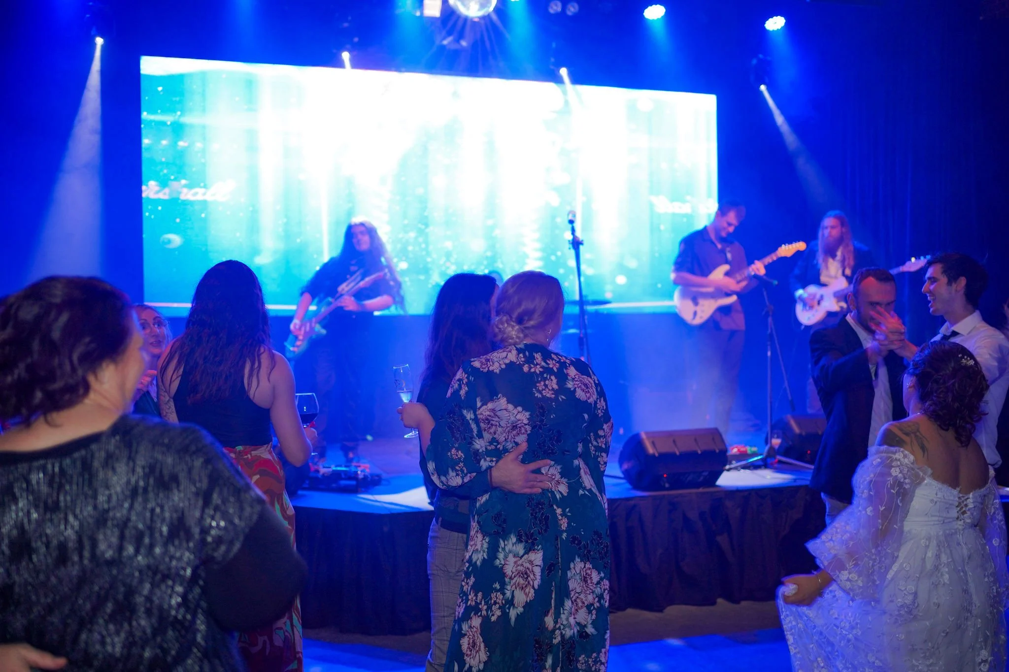 Wedding guests dancing near the stage while a live band plays under blue lighting and LED screens at Night Owl Studio.