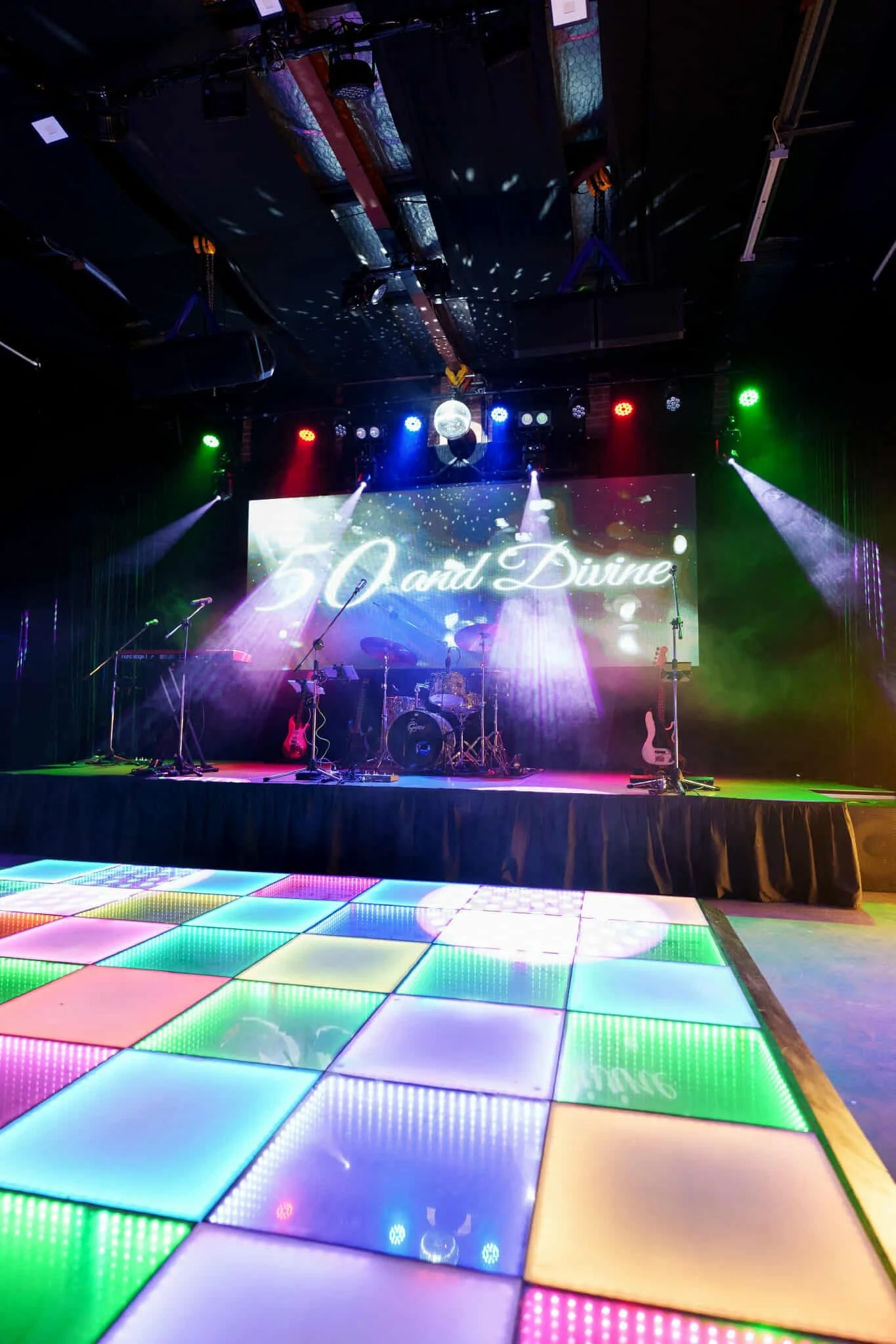 Front view of the colourful LED dance floor glowing in rainbow hues, leading up to a live music stage with instruments and a '50 and Divine' screen backdrop under disco lights