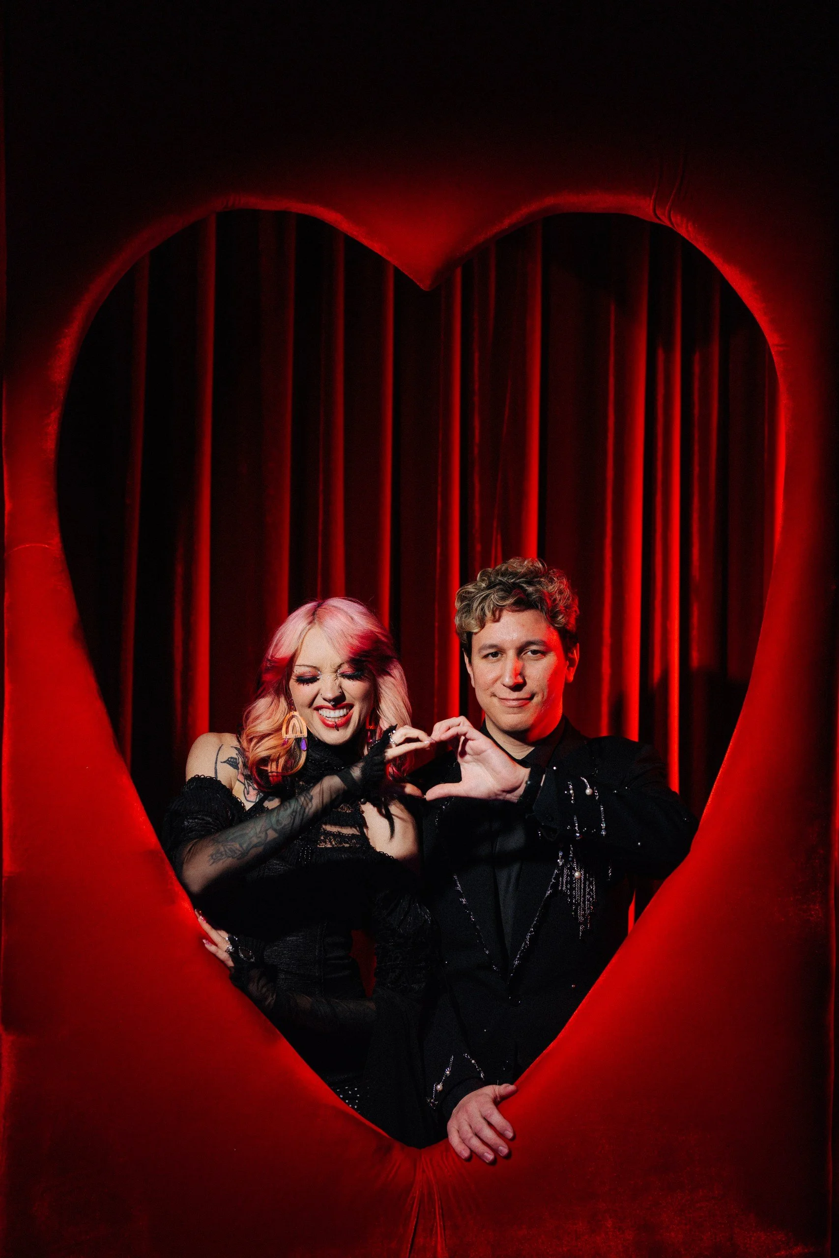 A couple poses together inside a large red heart against a red velvet curtain.