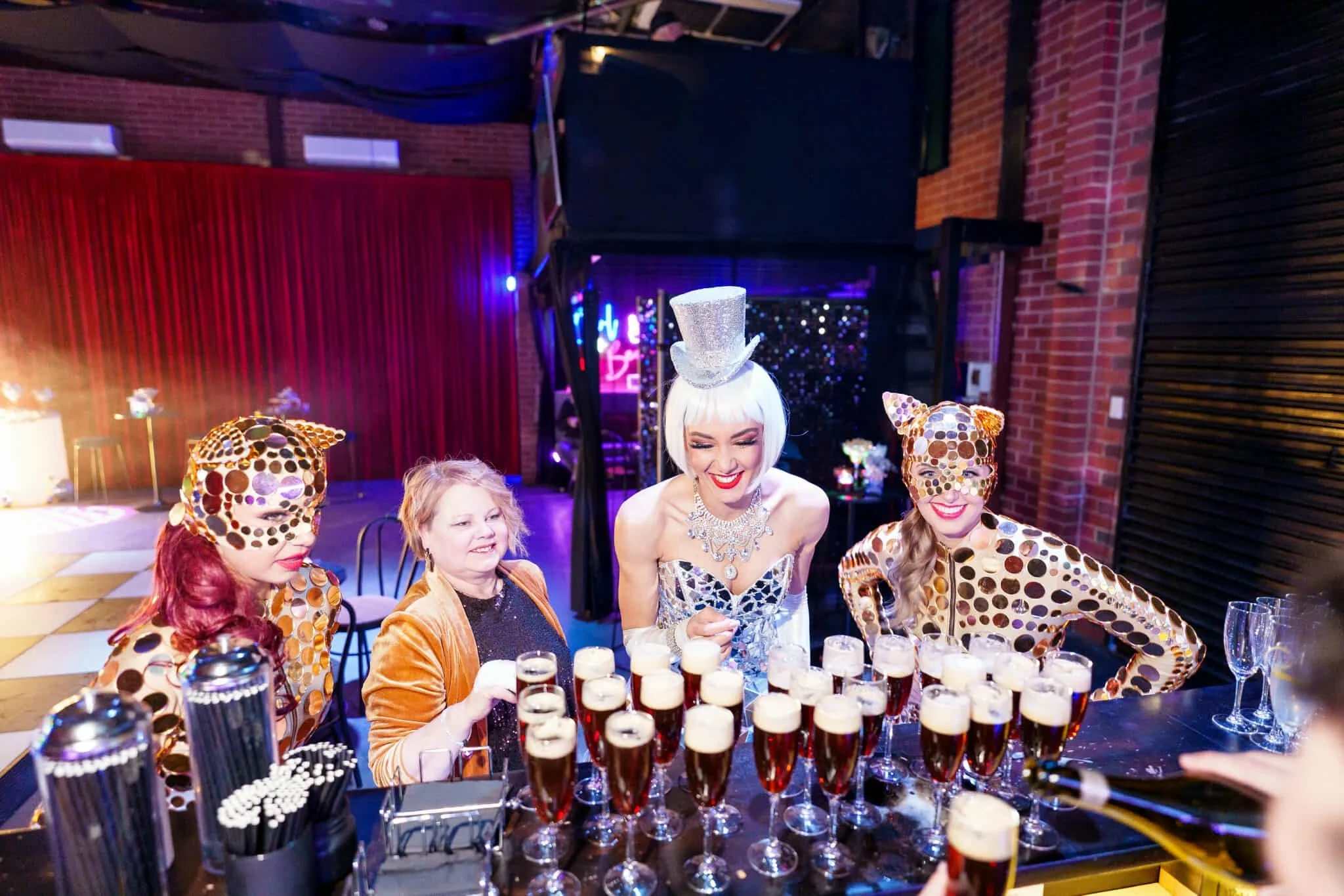 Performer in silver mirrorball corset and two gold leopard-suited dancers laughing with guests while serving cocktails at the bar.