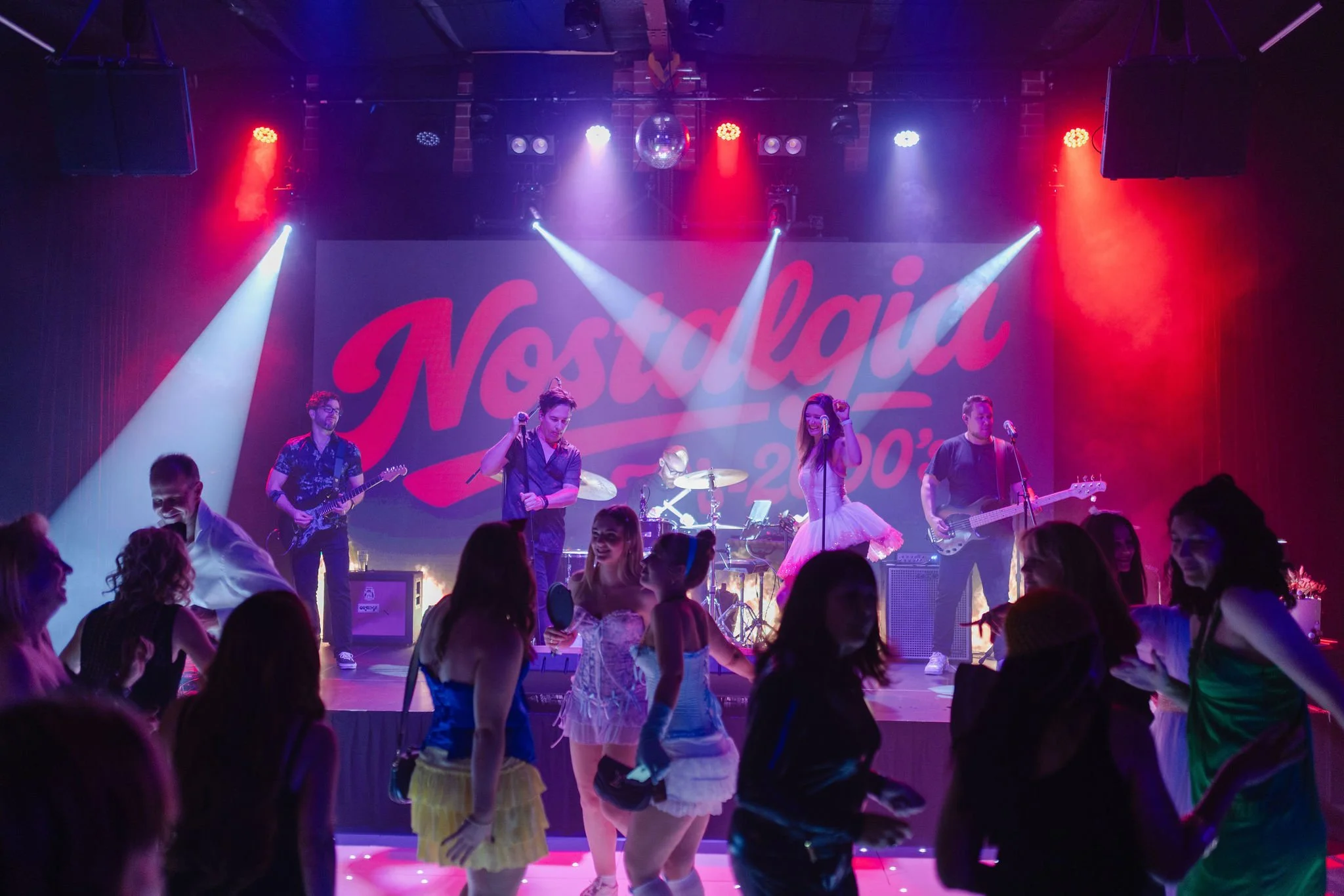 A wide shot of a crowded, dark venue with a band performing on a stage. The stage backdrop displays the word "Nostalgia" in large, retro-style red script with the text "2000's" underneath.