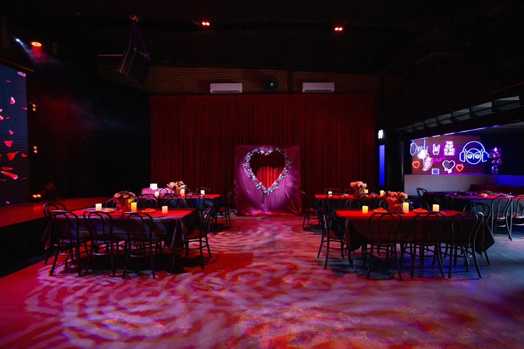 Long black tables with red runners and white candle centerpieces set up for an event. In the background, a large heart-shaped floral wreath stands in front of red curtains, next to a brick wall with neon signs.