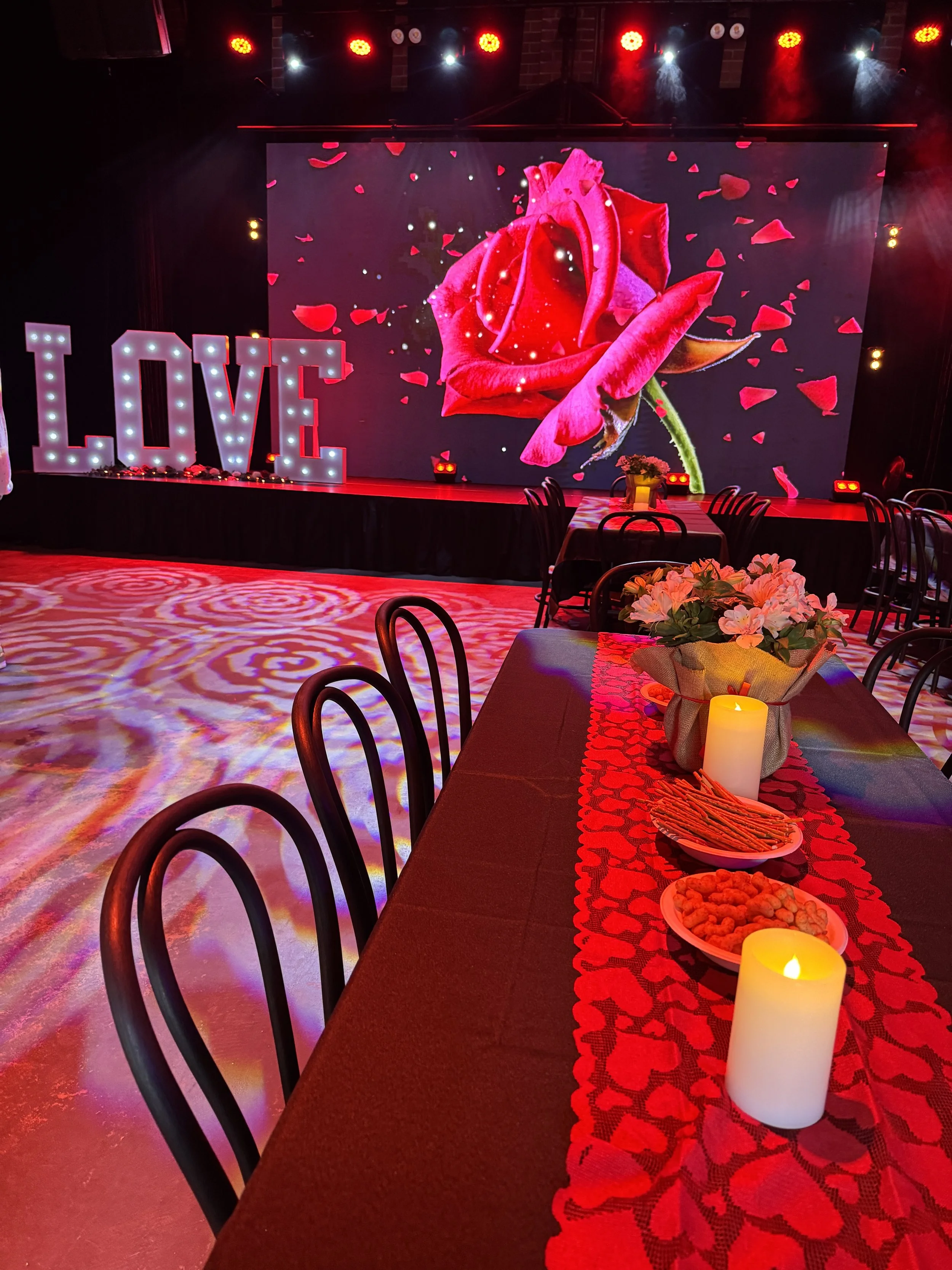 A close-up view of a banquet table dressed in a black tablecloth and a red heart-patterned runner, topped with lit pillar candles, snacks, and a floral centerpiece. In the background, black chairs sit on a floor illuminated with swirling red patterns