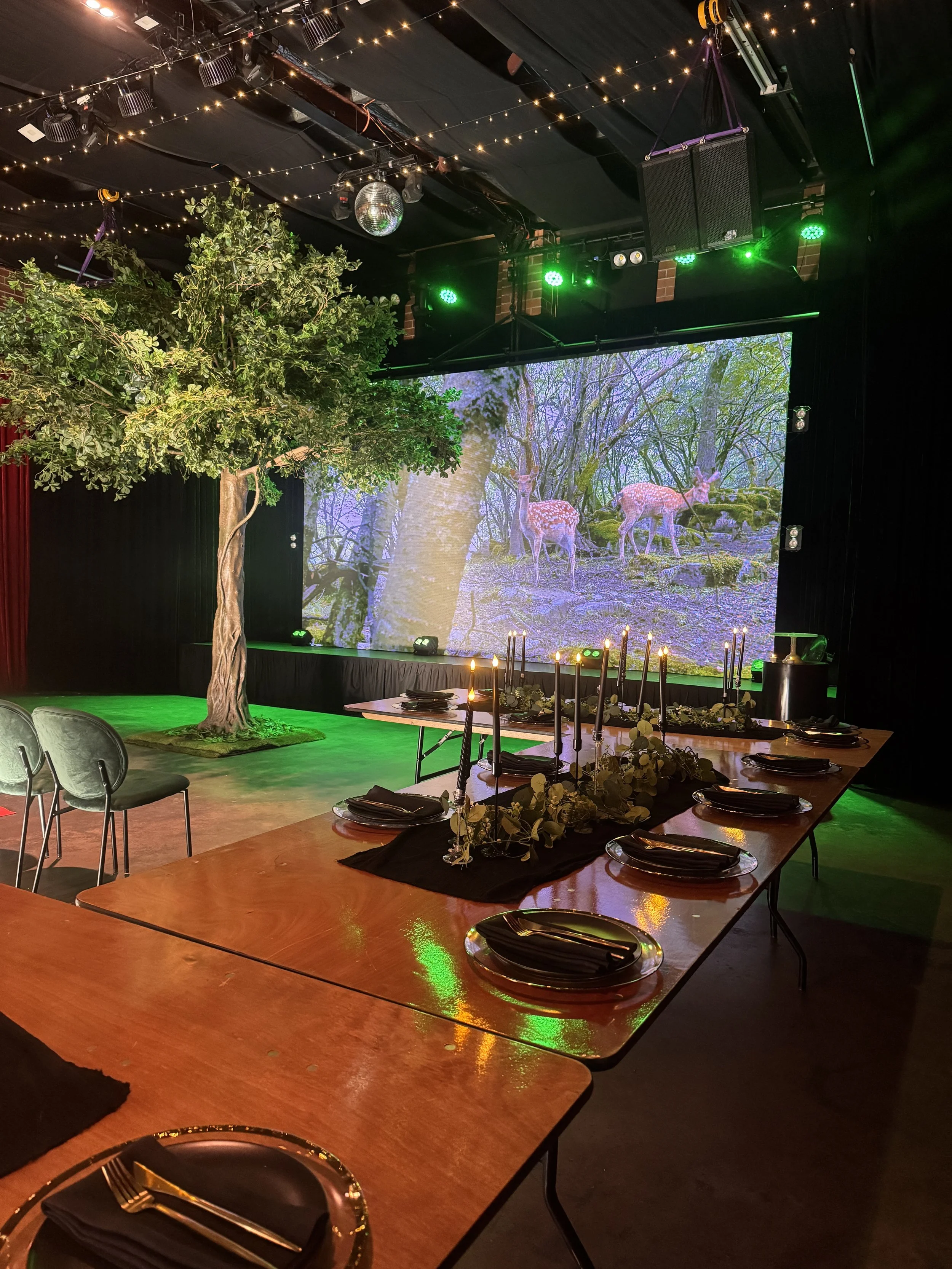 A high-angle view down a long wooden dining table set with black plates, gold cutlery, eucalyptus garlands, and tall black taper candles. In the background, a large tree prop and a forest-themed video screen create an enchanted atmosphere under ambie