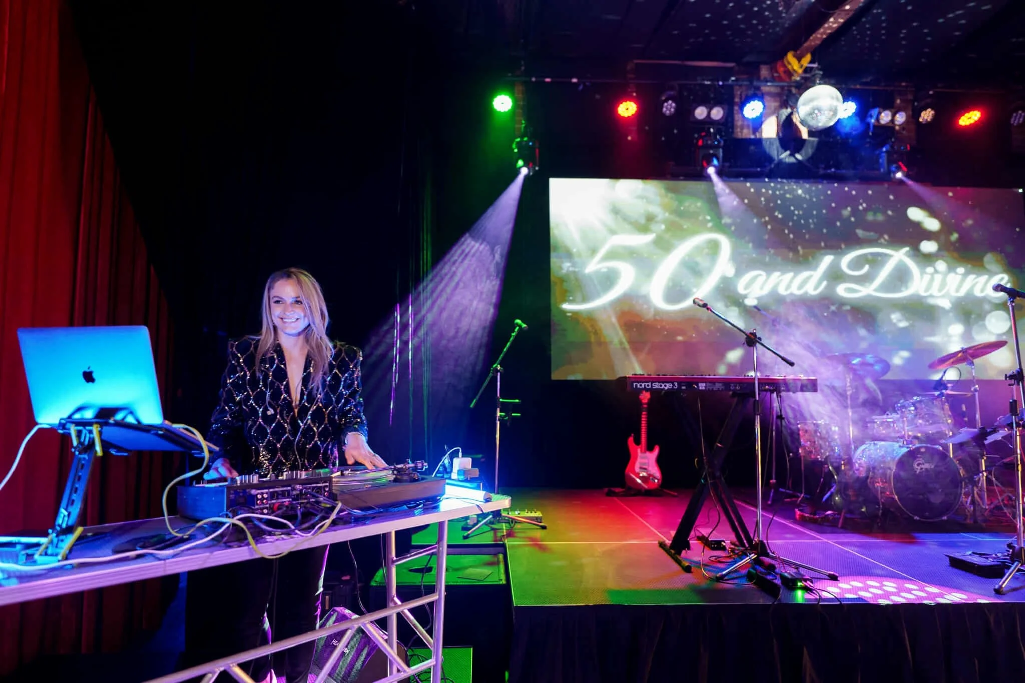 Female DJ smiling behind a modern setup with purple and green stage lights illuminating the instruments and '50 and Divine' screen behind her