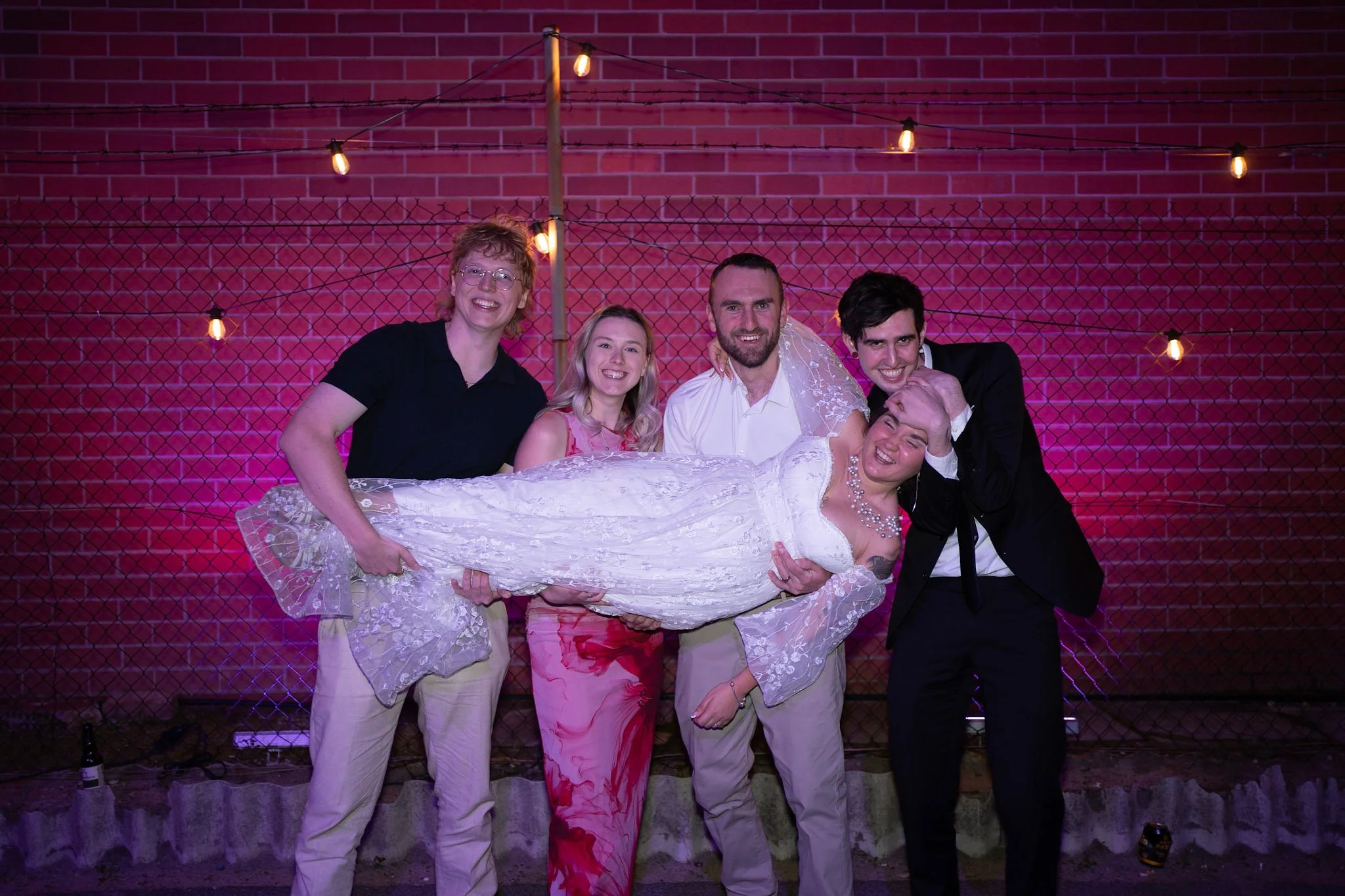 The bride is playfully held up by a group of friends in front of a brick wall strung with fairy lights, all smiling and laughing in a celebratory pose