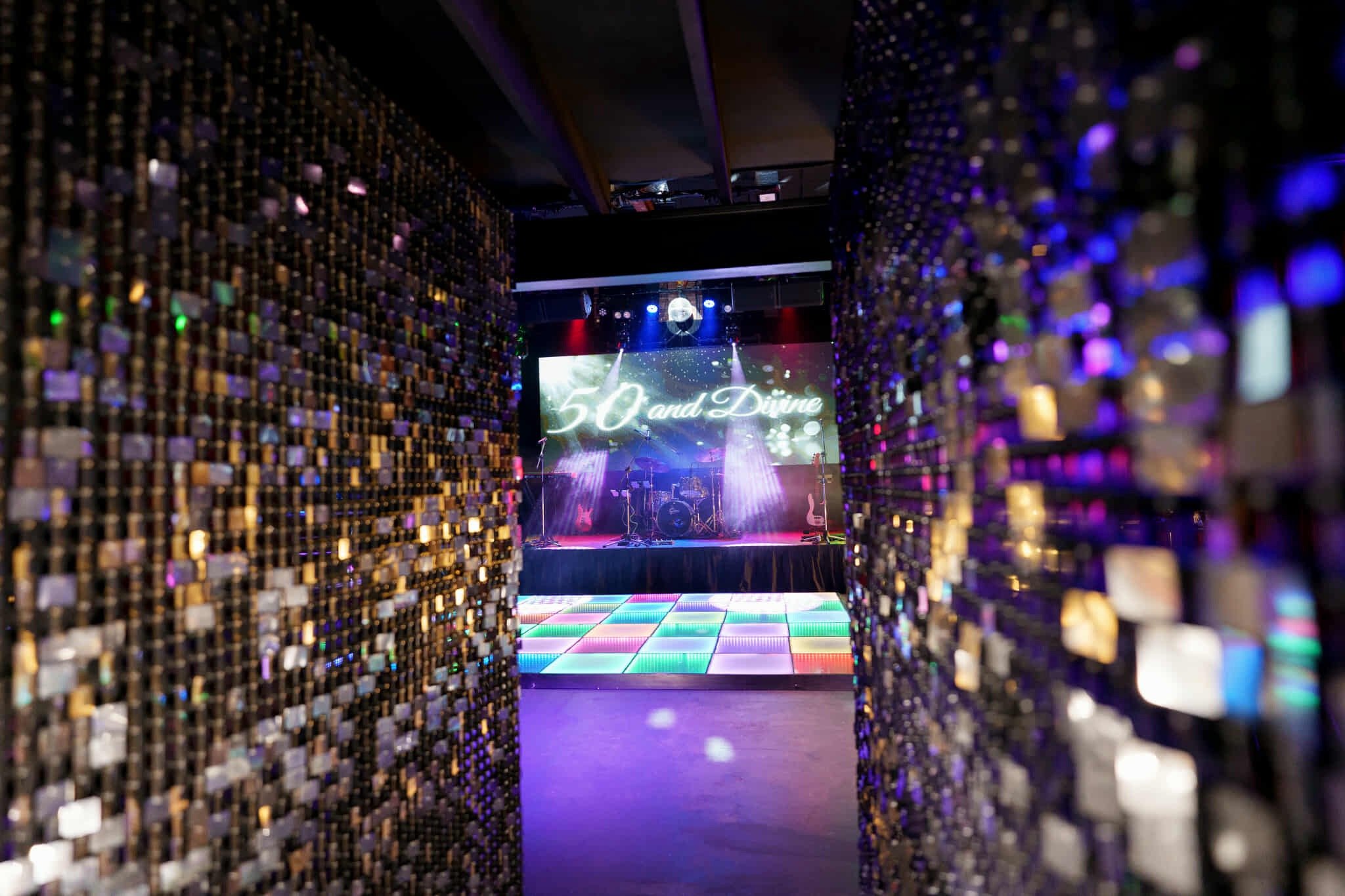 Glimpse through two glittering sequin feature walls revealing a stage setup with ‘50 and Divine’ projected on screen and a vibrant LED dance floor at Night Owl Studio.