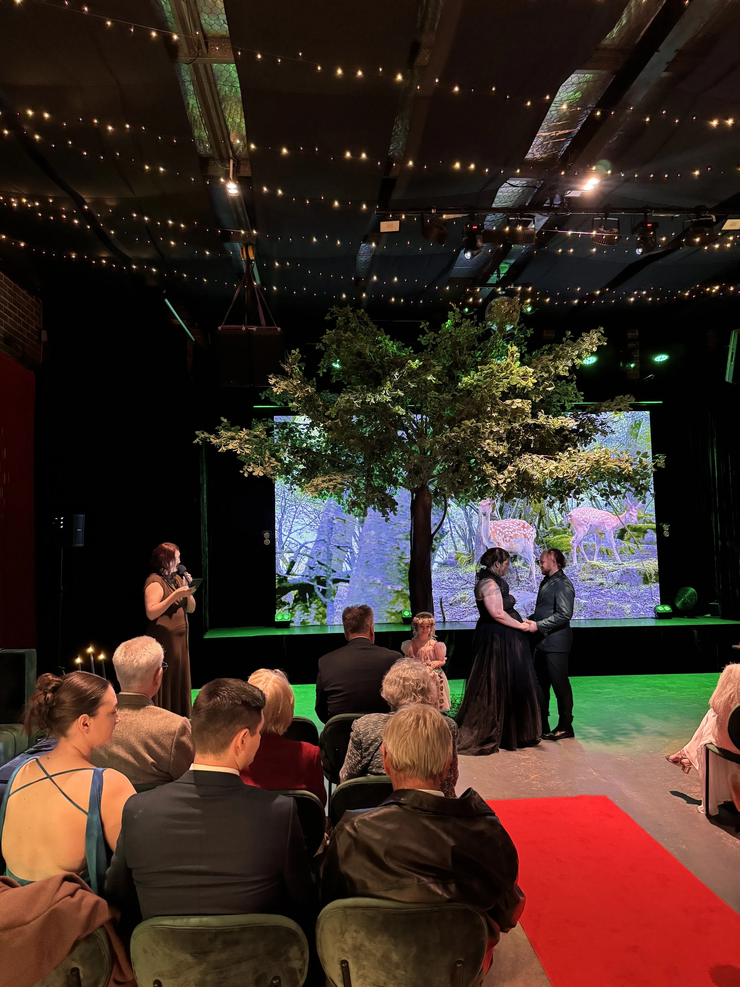 An audience seated in green velvet chairs watches a bride in a black dress and a groom in a dark suit stand together on stage. A large artificial tree stands beside them, illuminated by green stage lights against a screen displaying a forest landscap