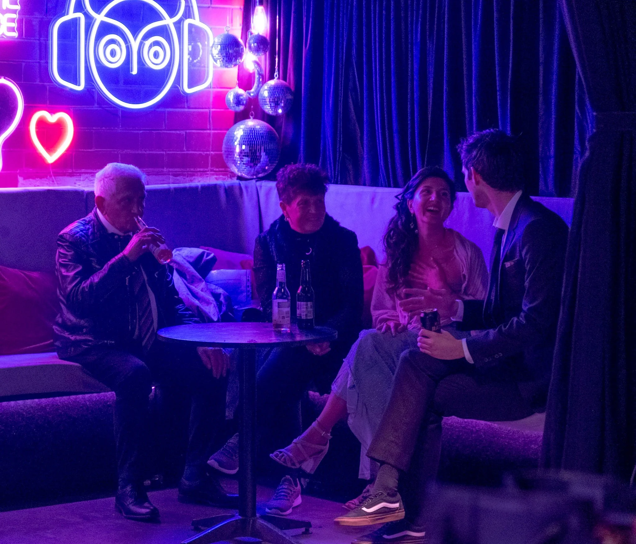 A group of guests sits relaxed in the neon-lit lounge corner of the venue, chatting and sipping drinks under glowing signs and disco balls.