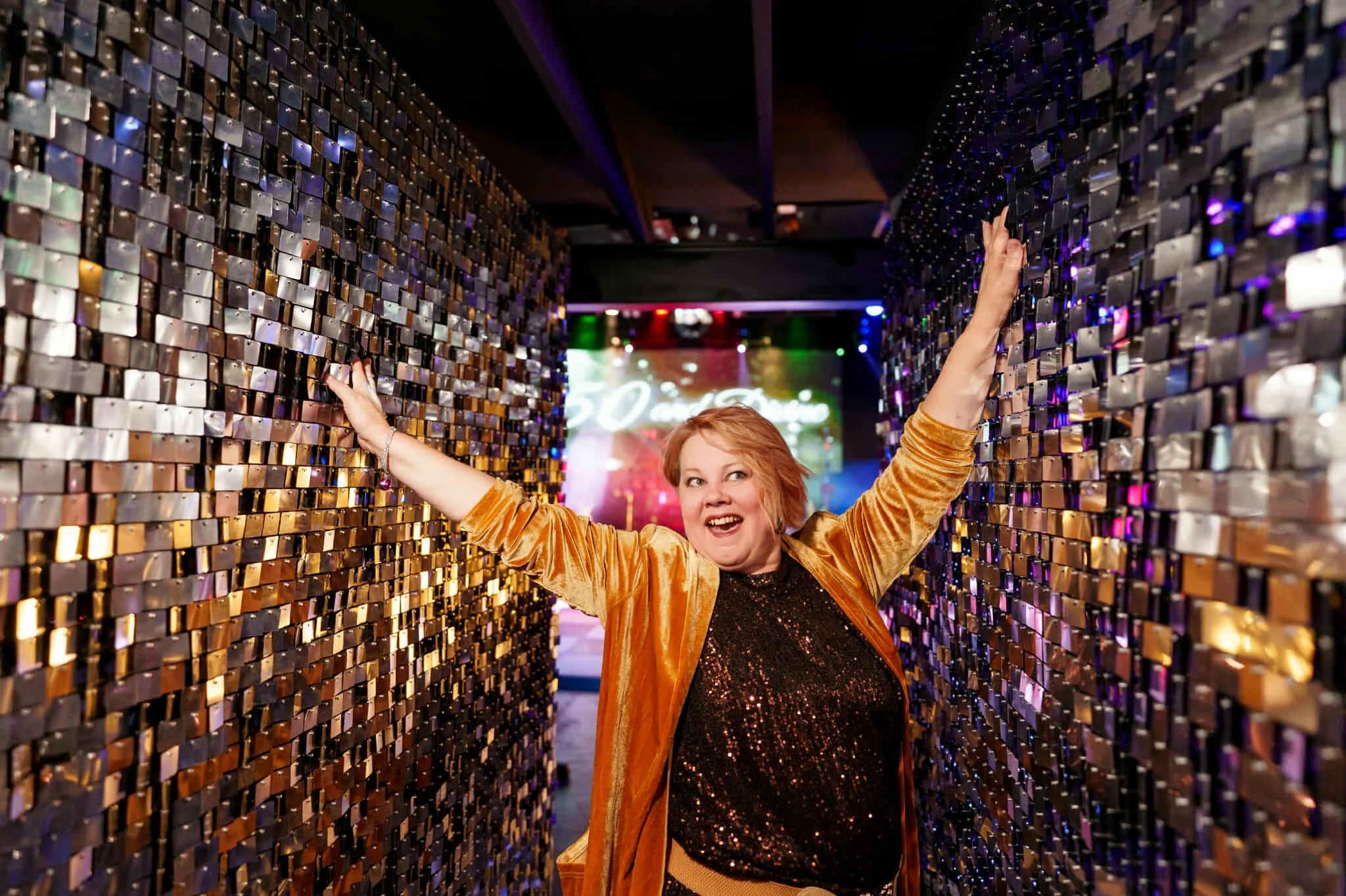 Rachel striking a joyful pose in a gold velvet jacket between two shimmering sequin feature walls at Night Owl Studio.