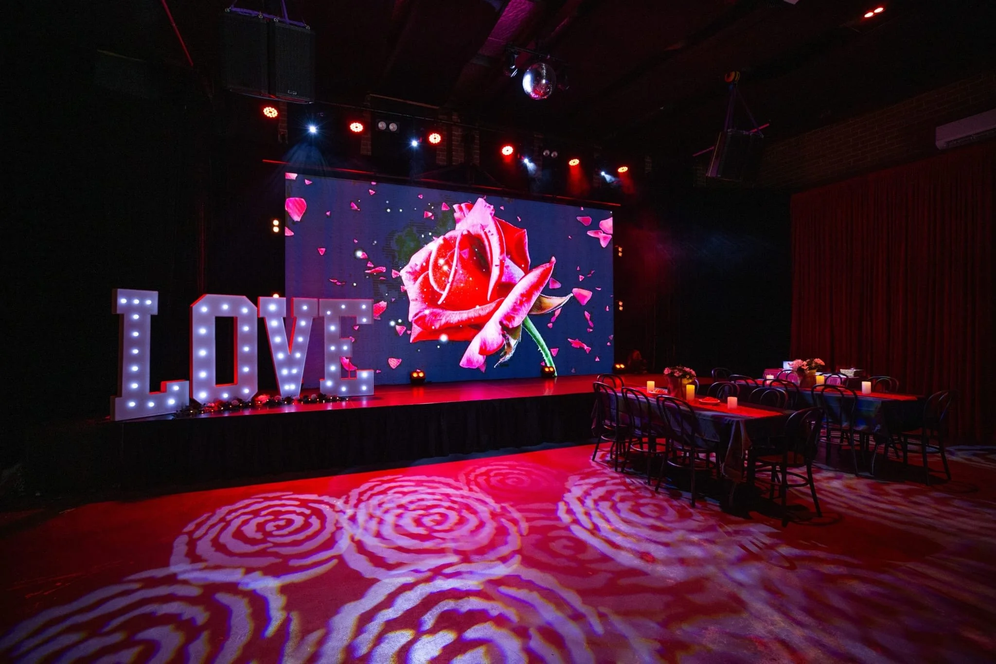 A romantic event space illuminated by large rose-patterned floor projections. Long tables with red heart runners lead toward a stage featuring large glowing "LOVE" letters and a massive screen displaying a red rose.