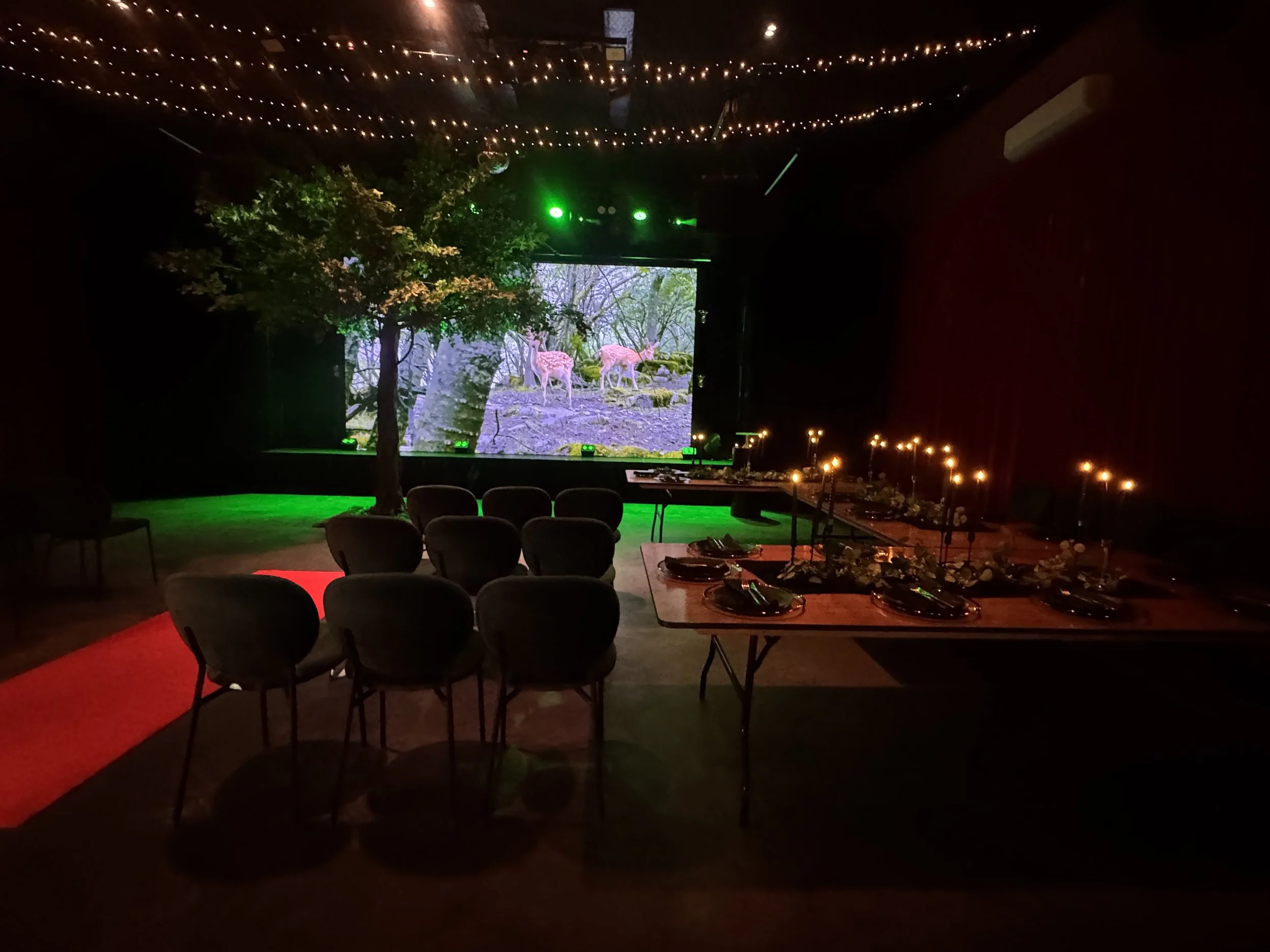 A dimly lit venue room featuring a long banquet table set with black tableware, greenery, and glowing taper candles. In the background, green stage lighting illuminates a large leafy tree standing beside a screen showing a peaceful forest scene.