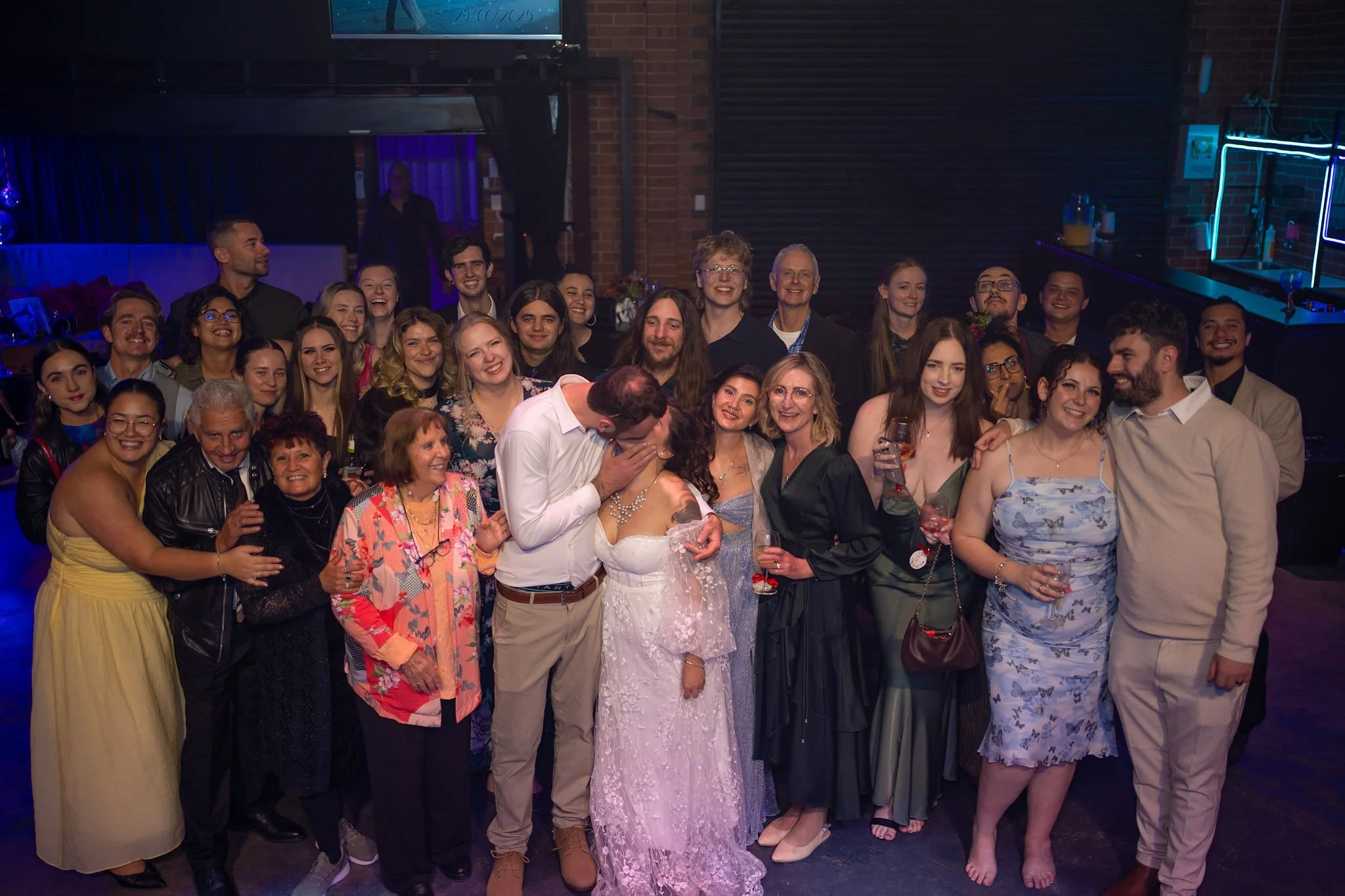 Large group photo of guests gathered around the bride and groom, smiling, hugging, and celebrating the night in a lively studio setting.