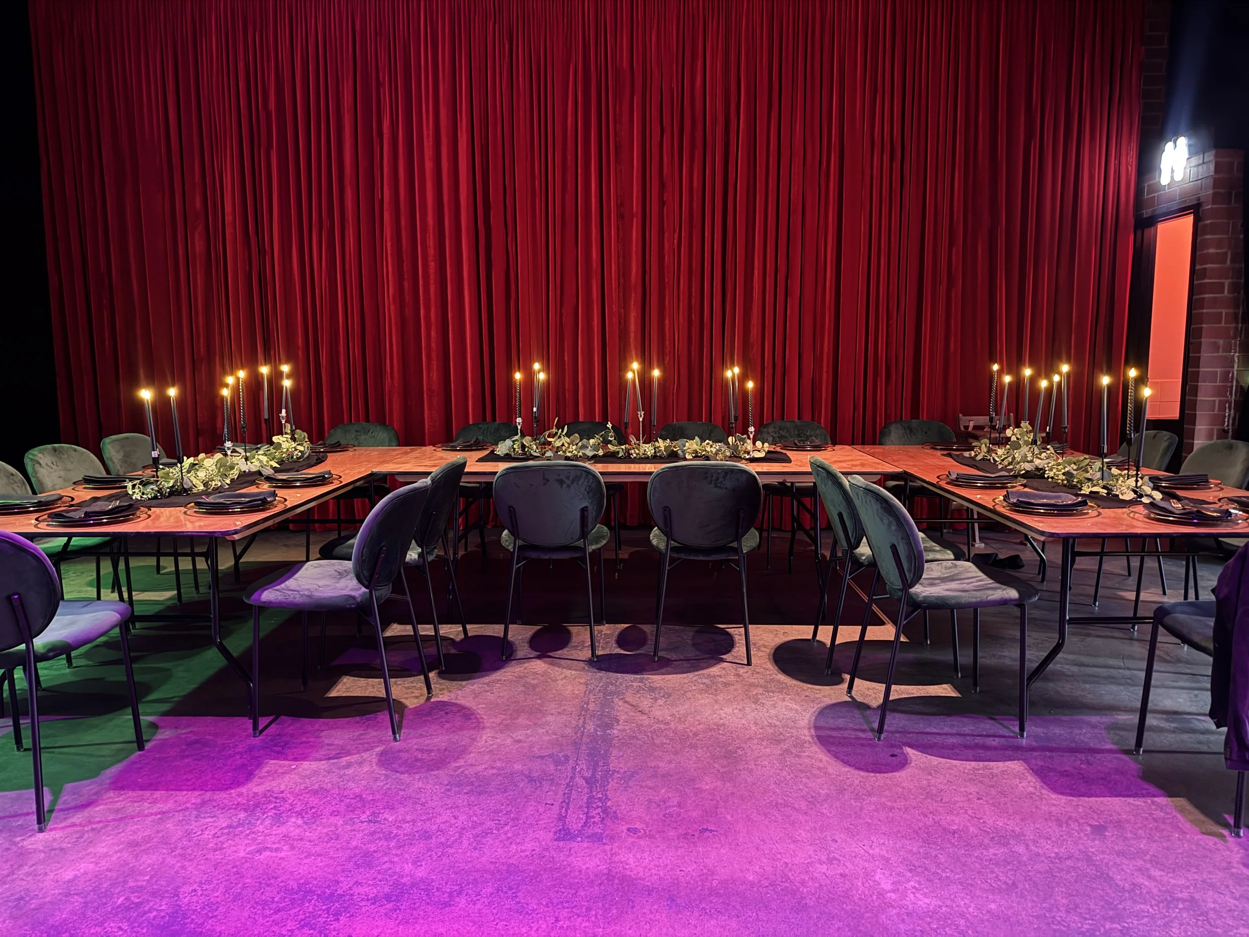 Long wooden banquet tables arranged in a U-shape setup in front of floor-to-ceiling red velvet curtains. Dark green velvet chairs surround the tables, which are decorated with eucalyptus runners and numerous tall taper candles glowing under magenta f