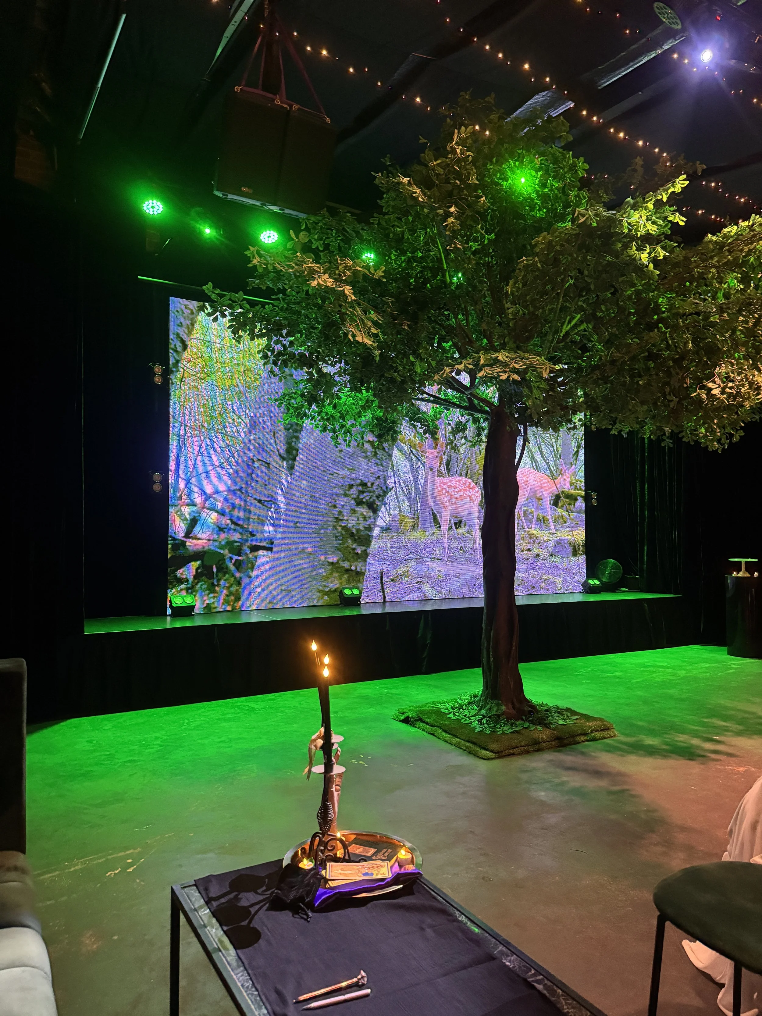 A close-up view of the indoor tree prop lit with vibrant green stage lighting. A small table in the foreground holds a warm candle display alongside a stage backdrop displaying a detailed forest scene.