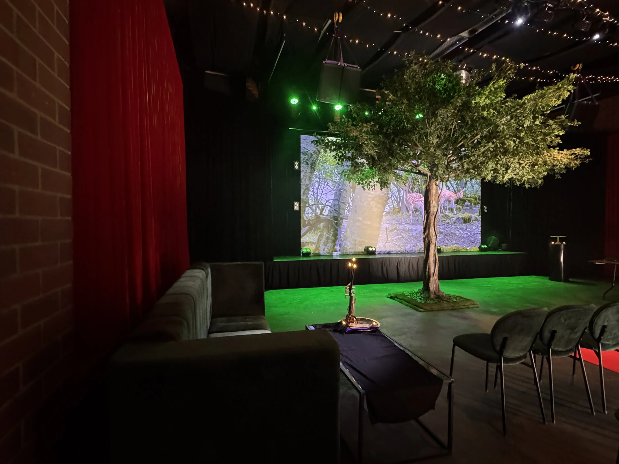 A dark green velvet couch and matching chairs frame a low coffee table decorated with a lit candle. In the background, an artificial tree stands under green spotlights in front of a stage screen displaying a woodland forest scene.