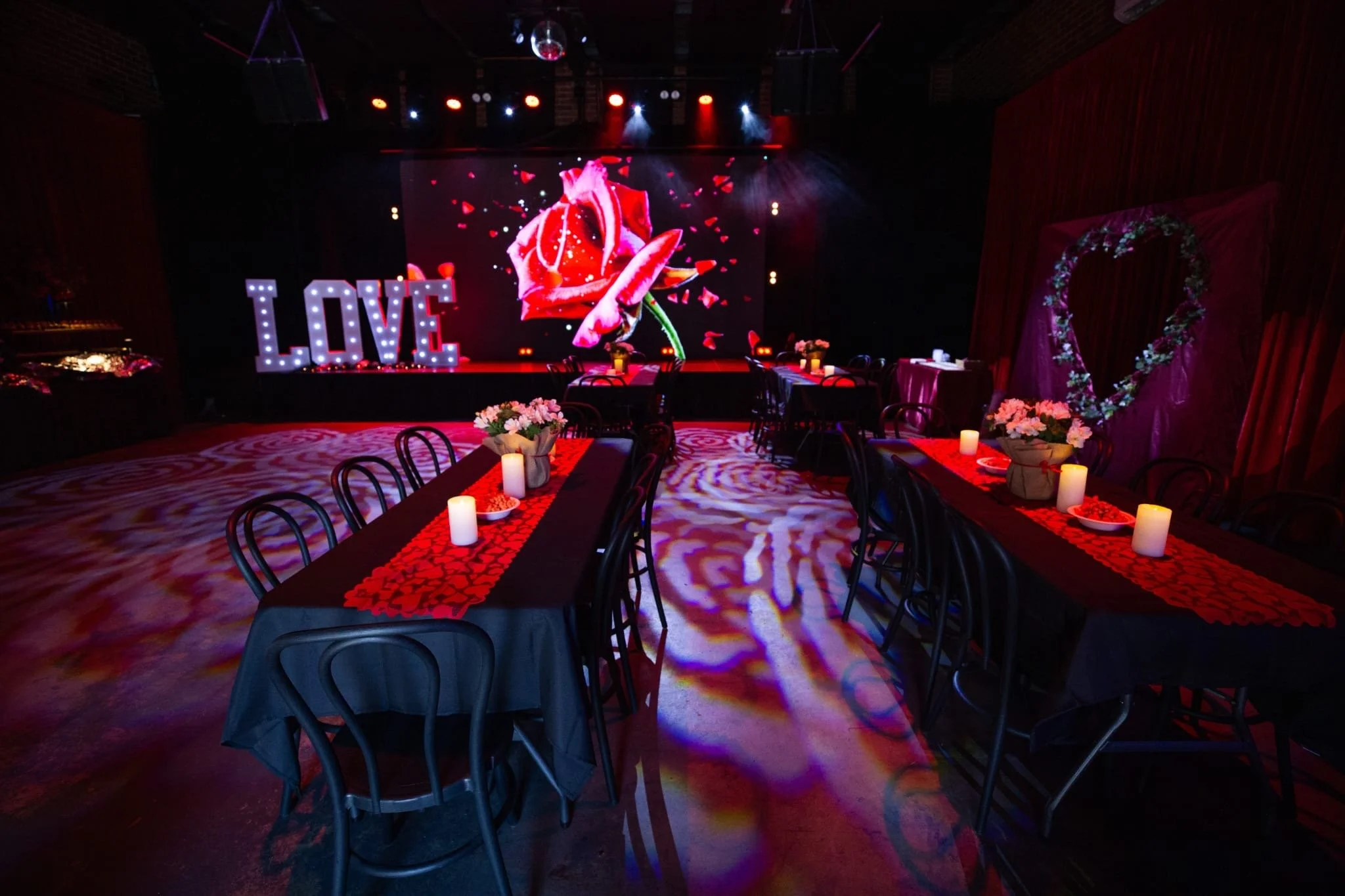 A wide view of a romantic event setting showing dining tables in the foreground. The background features a stage with illuminated "LOVE" letters and a large screen displaying a rose on the left, and a floral heart backdrop standing in front of red cu