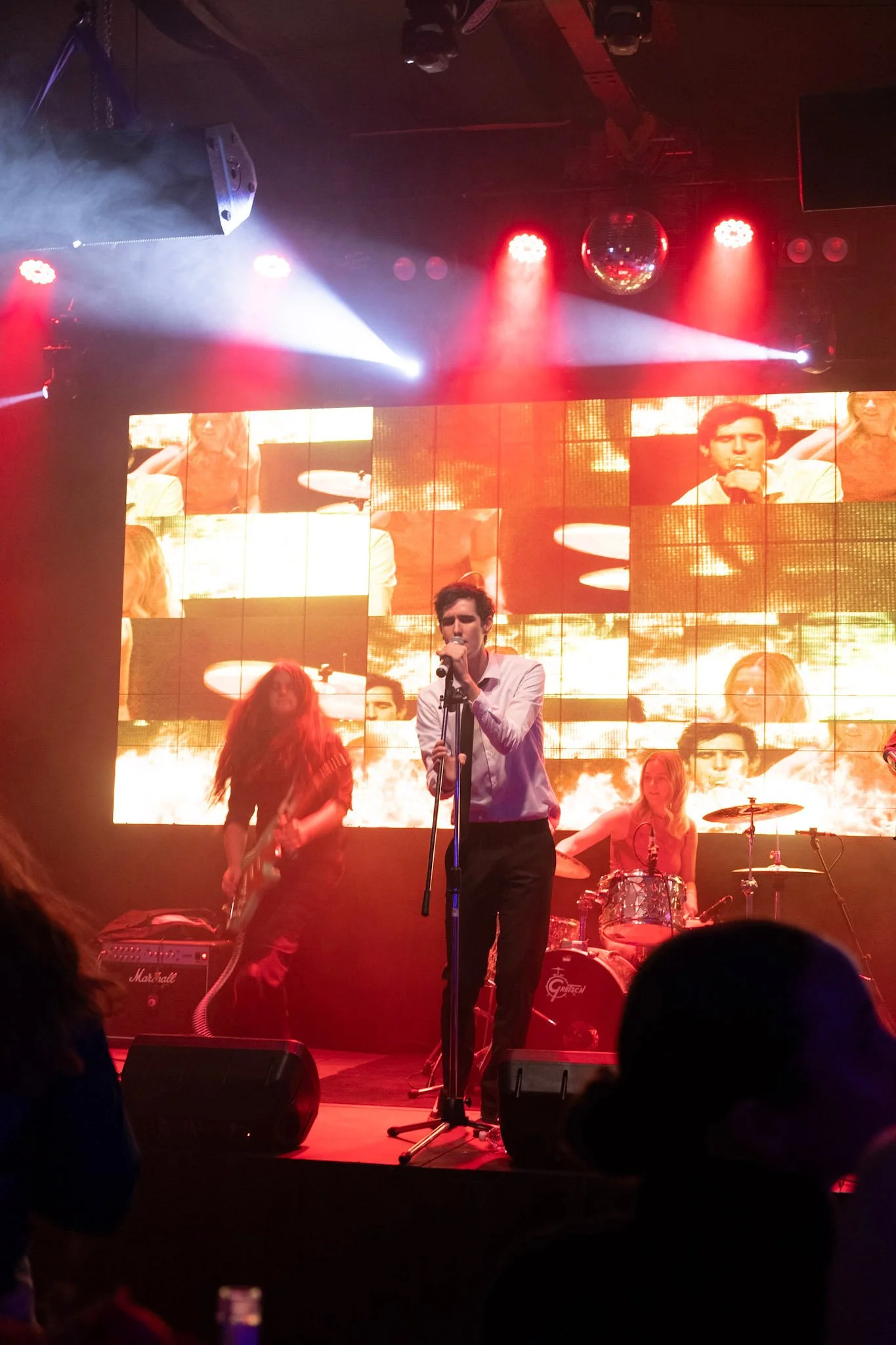 The lead singer performs center stage in a white shirt and black tie, surrounded by dramatic lighting and mirrored visuals on a glowing LED screen.