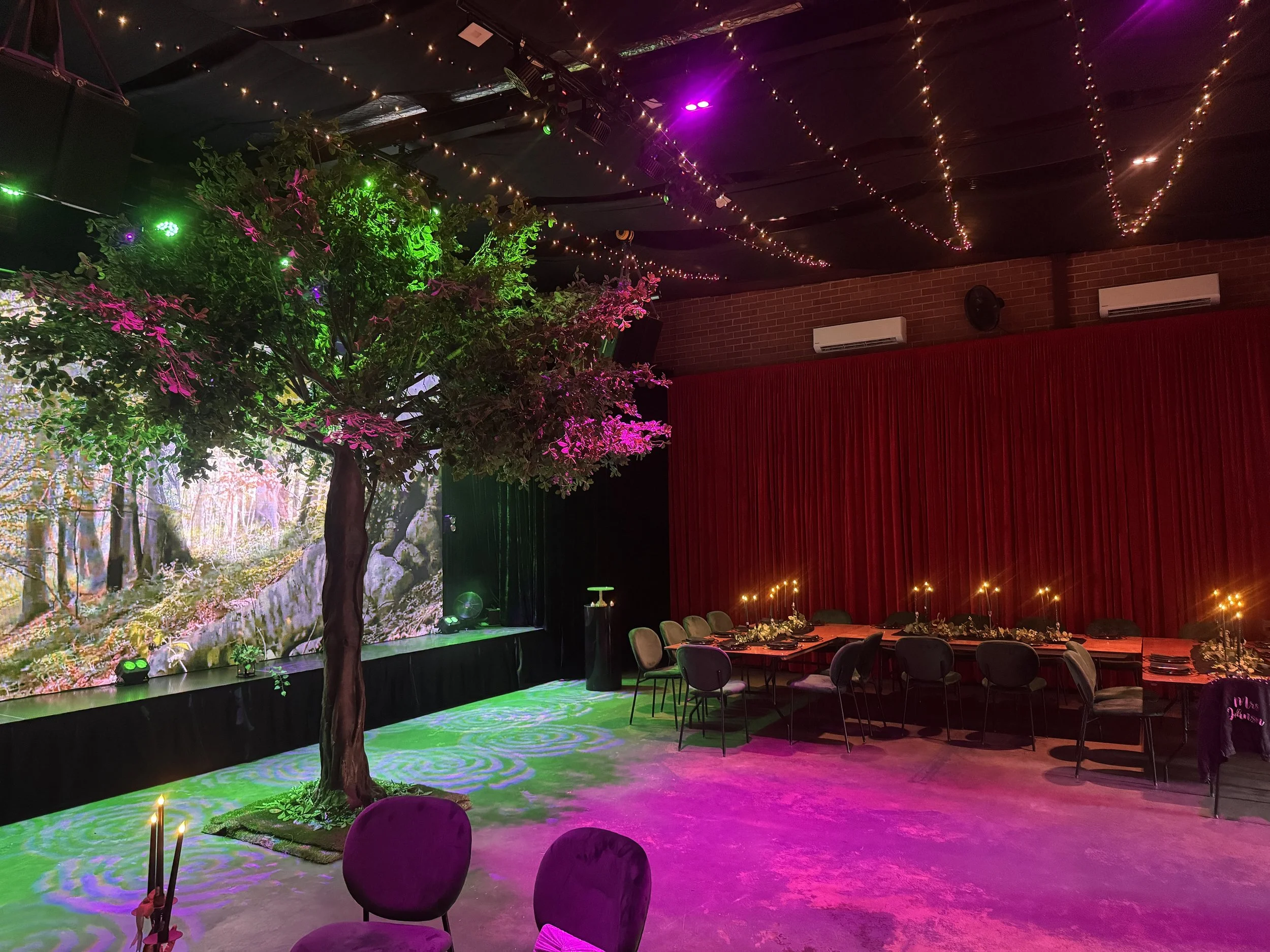 A broad view of an indoor event venue styled with an enchanted forest theme. An artificial tree bathed in green and pink lighting stands beside a stage screen showing a woodland scene, while long banquet tables with glowing candles and velvet chairs 