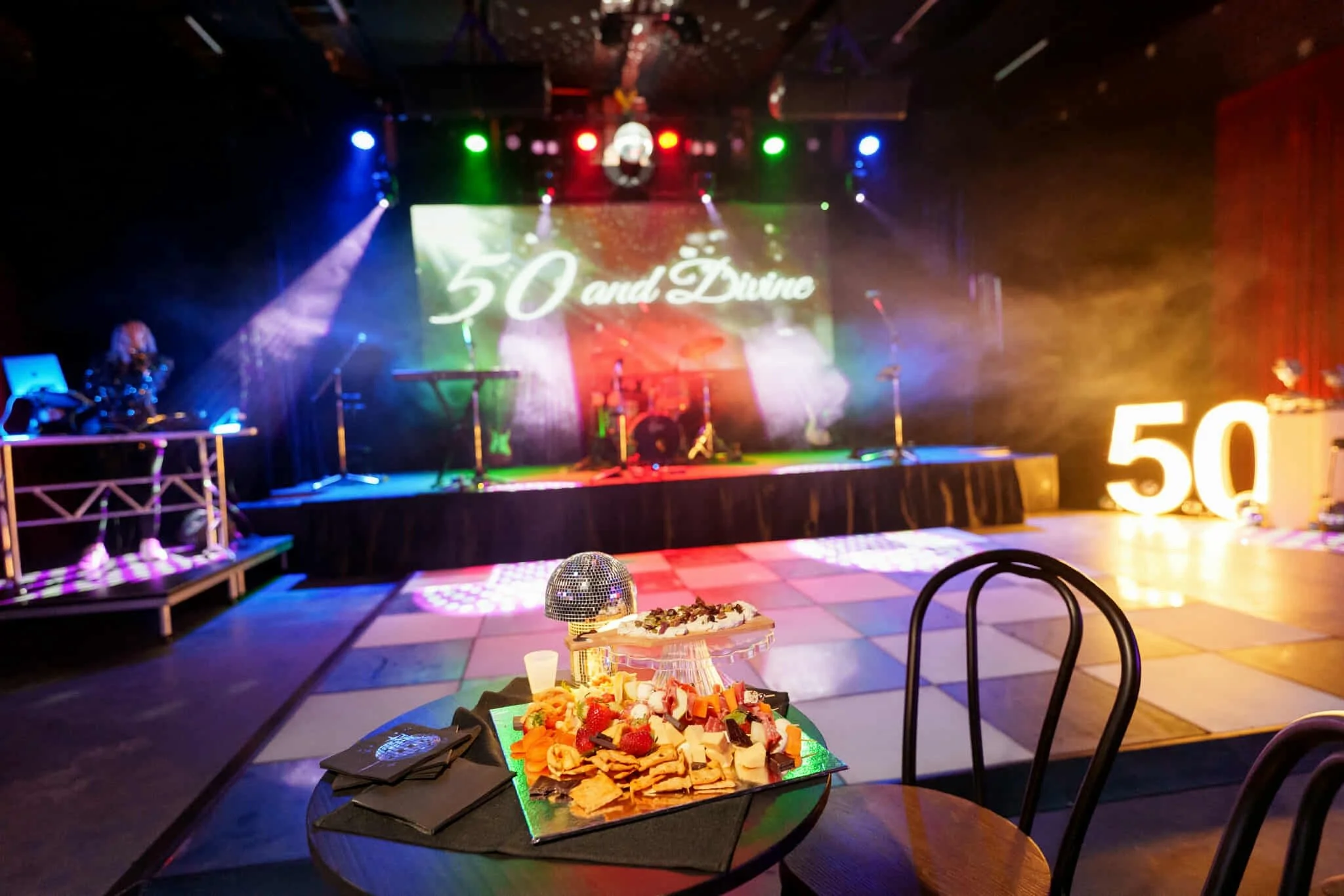 Grazing platter on a cocktail table in front of the lit stage and '50' light-up numbers, with a DJ playing in the background.