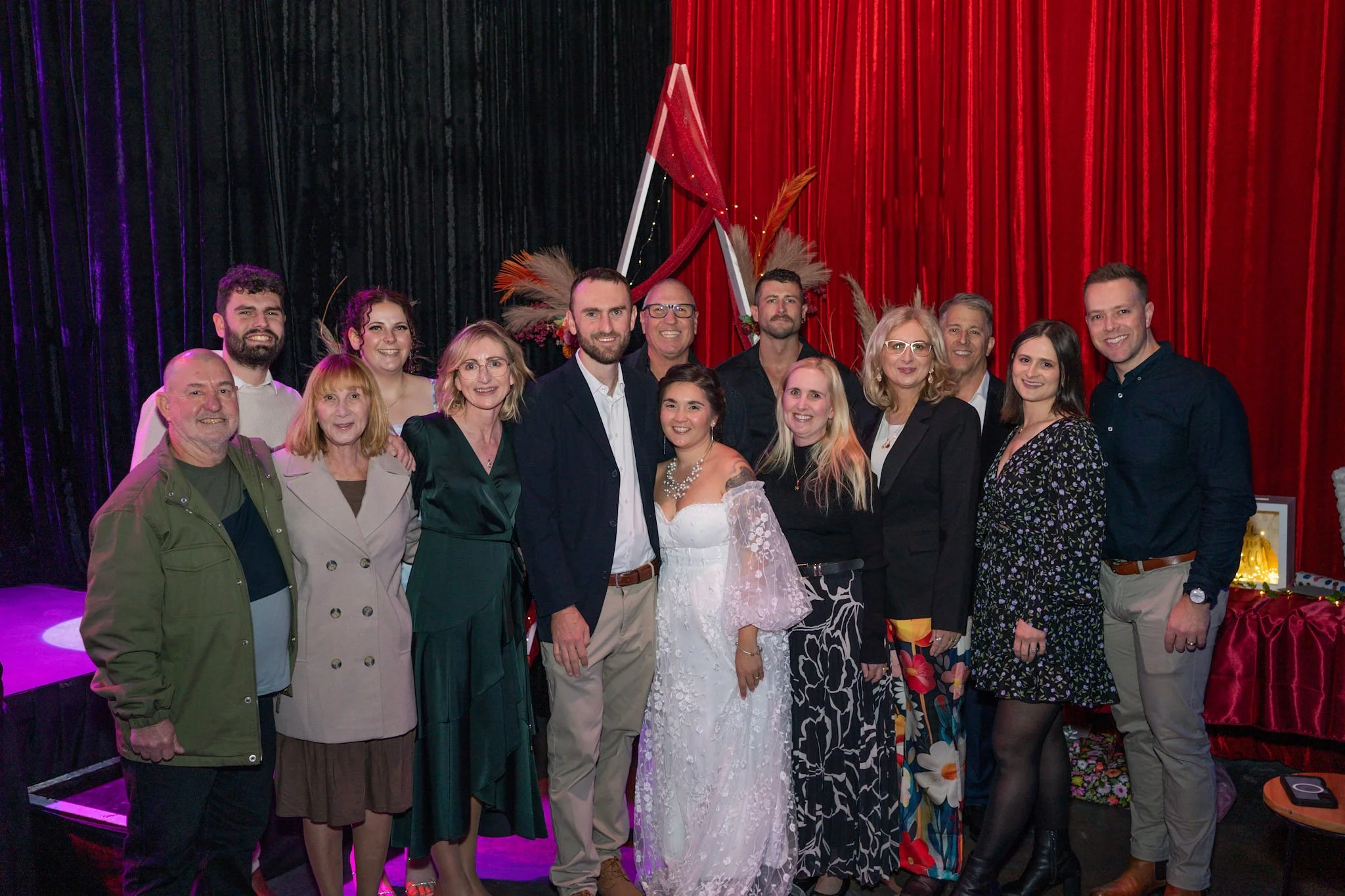 Yanina and Alex surrounded by friends and family in front of a red velvet curtain and triangle floral arbour at Night Owl Studio