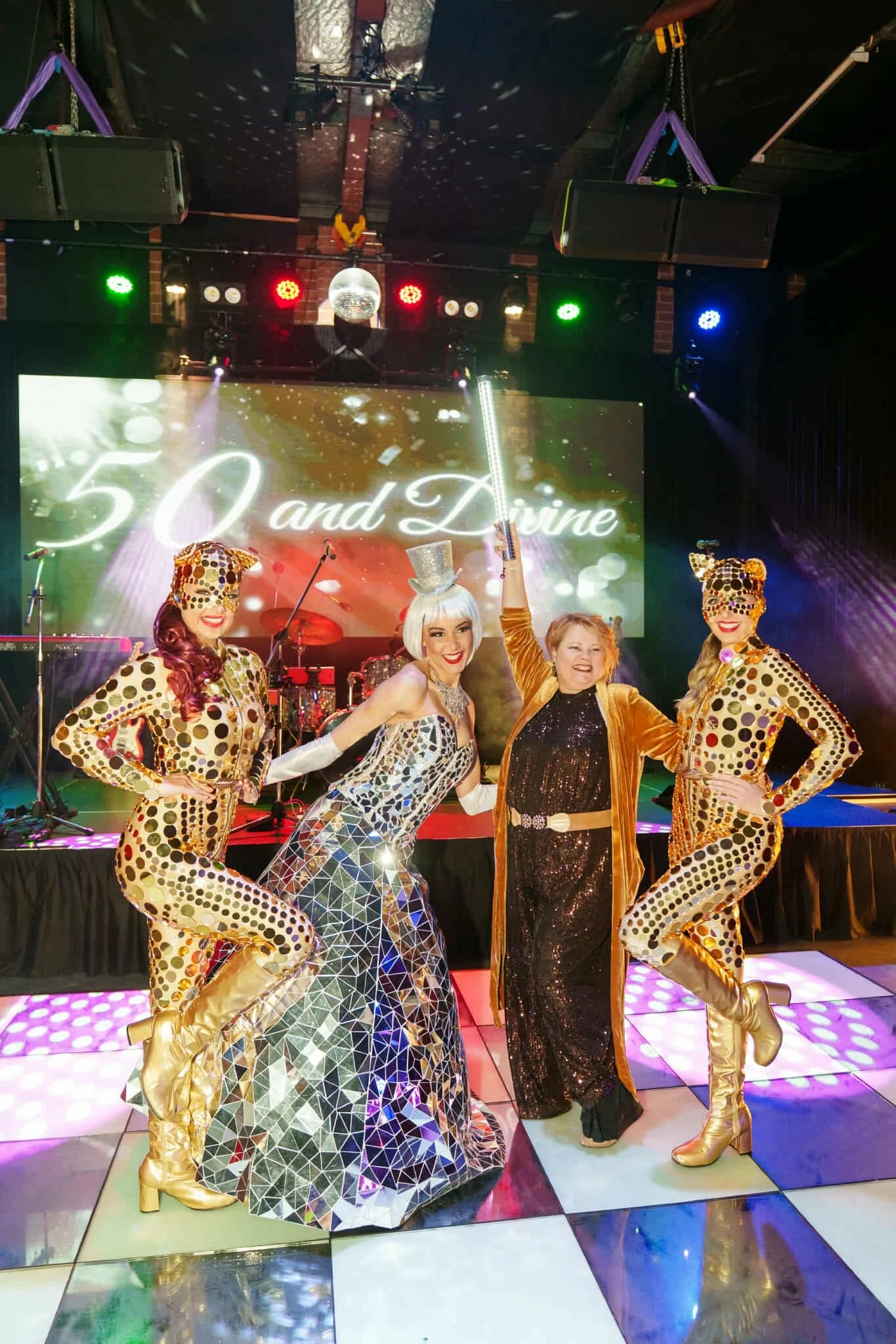 Rachel posing on a checkered dance floor with performers dressed in gold and mirrorball outfits, in front of a large ‘50 and Divine’ screen.