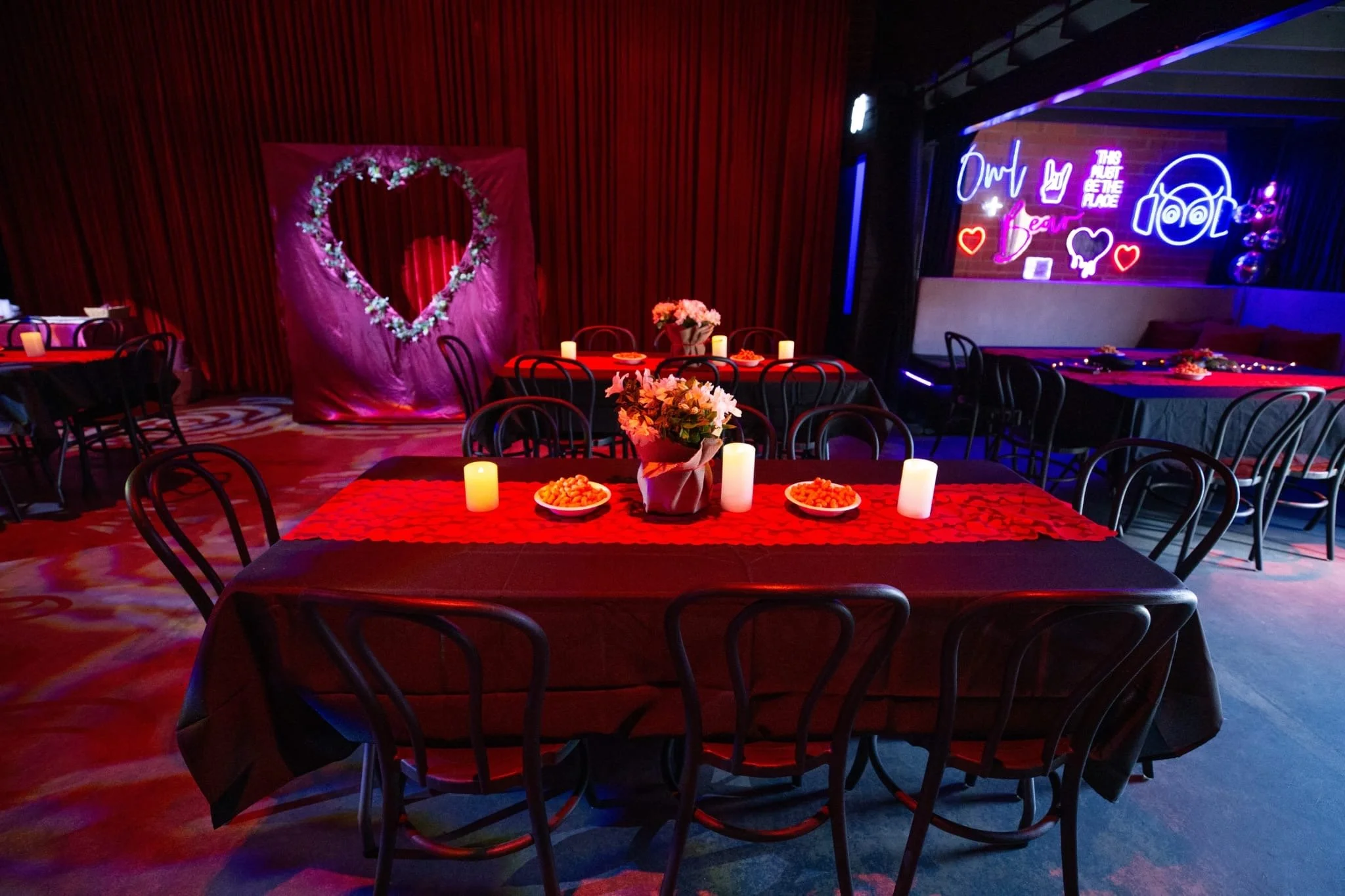 An event space with long dining tables set under red lighting. In the background, there is a large heart-shaped floral photo backdrop against red curtains and a neon sign on a brick wall that reads "Owl Bear" and "This Must Be The Place".