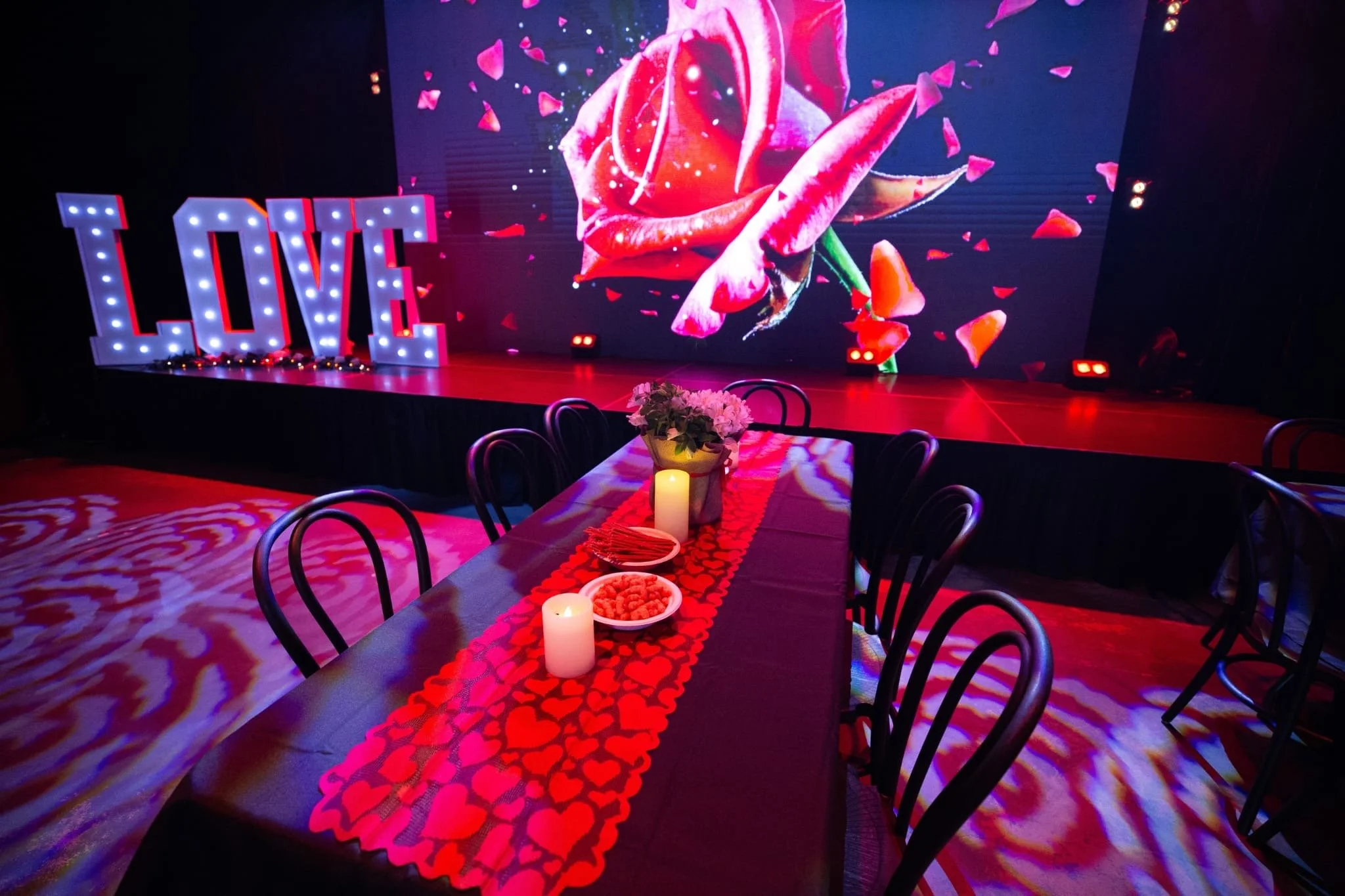 A brightly lit stage area featuring large marquee letters spelling "LOVE" in front of a large LED screen showing a red rose with falling petals. The venue floor is heavily covered in red and purple rose-patterned lighting.