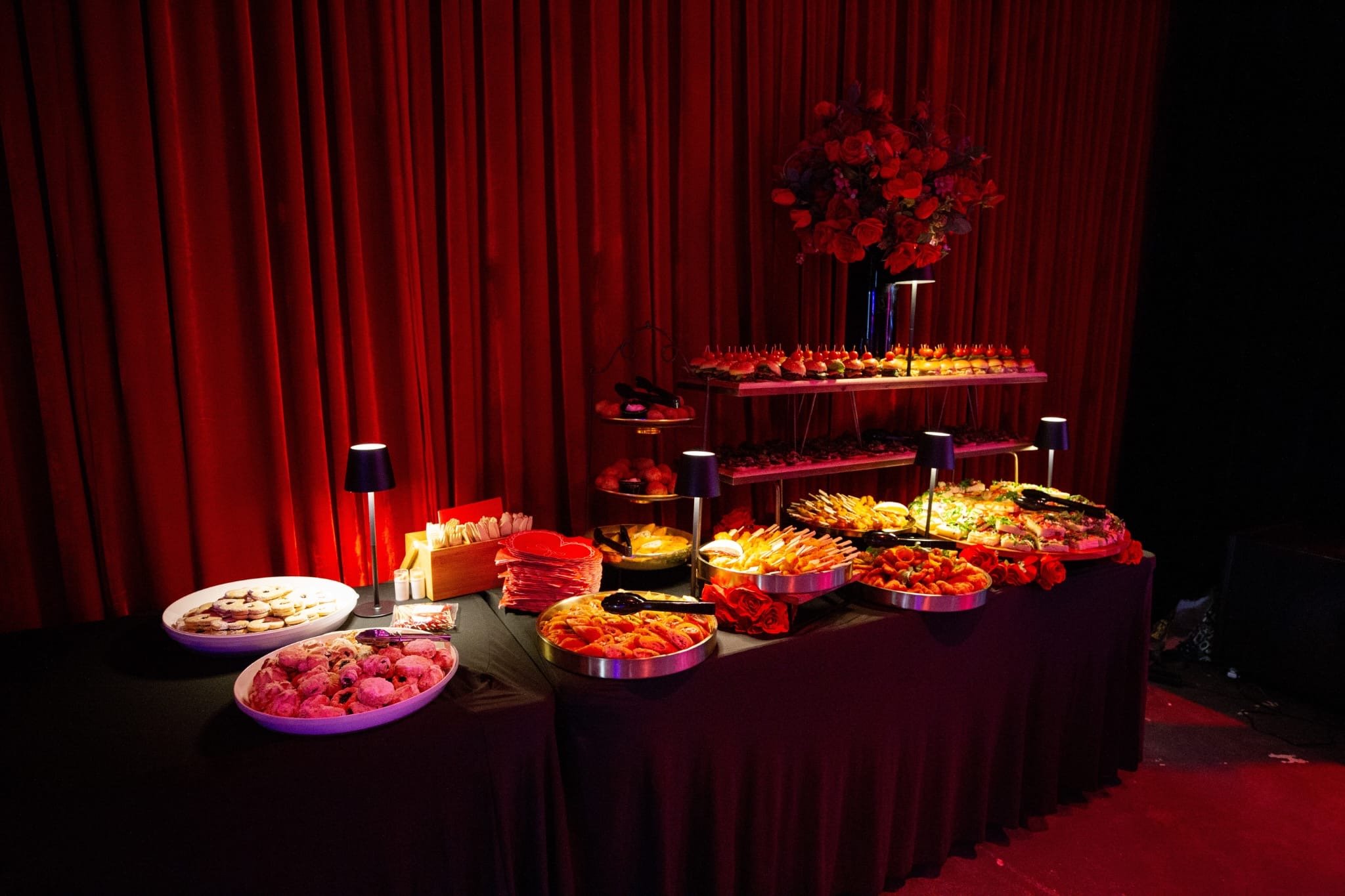 An angled view of a catering buffet table with a black tablecloth and red curtain backdrop. The spread includes tiered stands of mini burgers alongside platters of sandwiches, skewers, and desserts.