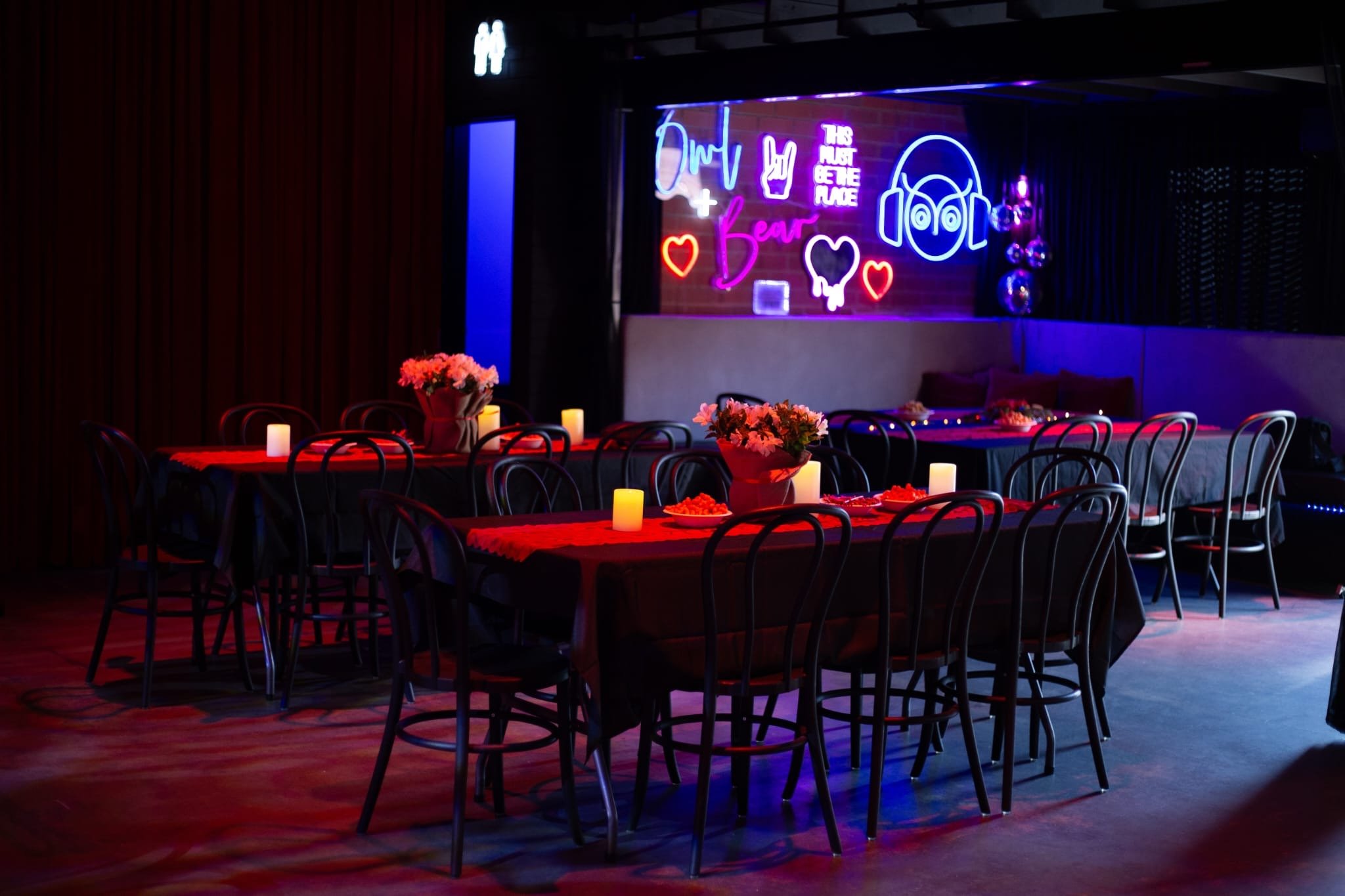 A dining area in a dimly lit venue with a long table decorated with a red runner, candles, and a floral centerpiece. Behind the seating area, a neon sign featuring an owl wearing headphones and the words "Owl Bear" glows on a brick wall next to disco