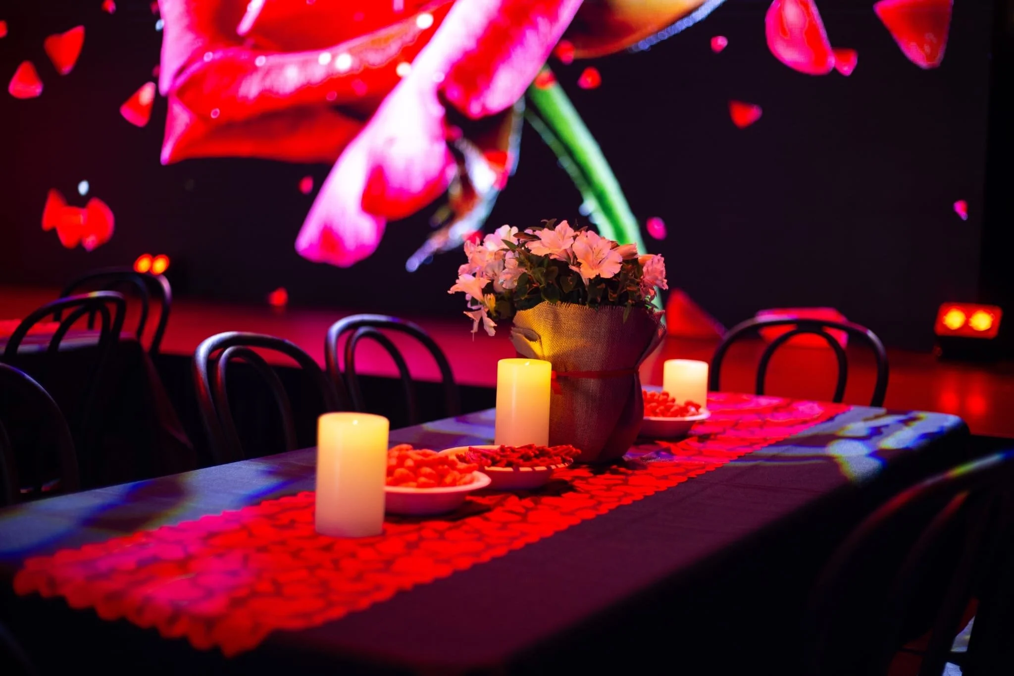 A close-up of a banquet table set with a red heart-cutout table runner, white pillar candles, and a floral arrangement. In the background, a stage features glowing "LOVE" letters and a large screen displaying a blooming red rose.