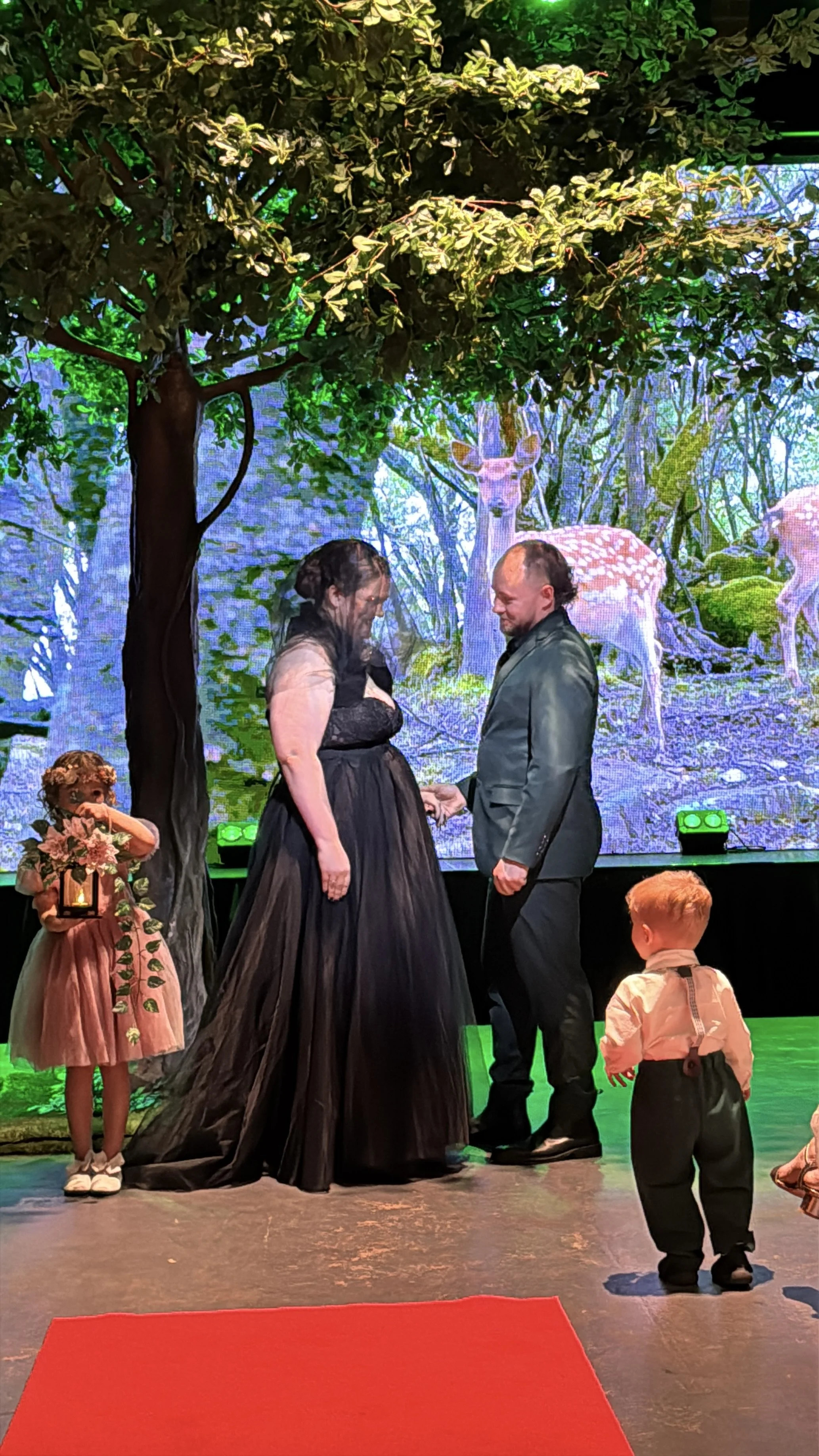 A bride in an off-the-shoulder black gown and veil holds hands with her groom in a dark green suit during their ceremony. They stand under the branches of an artificial tree on stage in front of a forest screen, while two young children stand nearby.