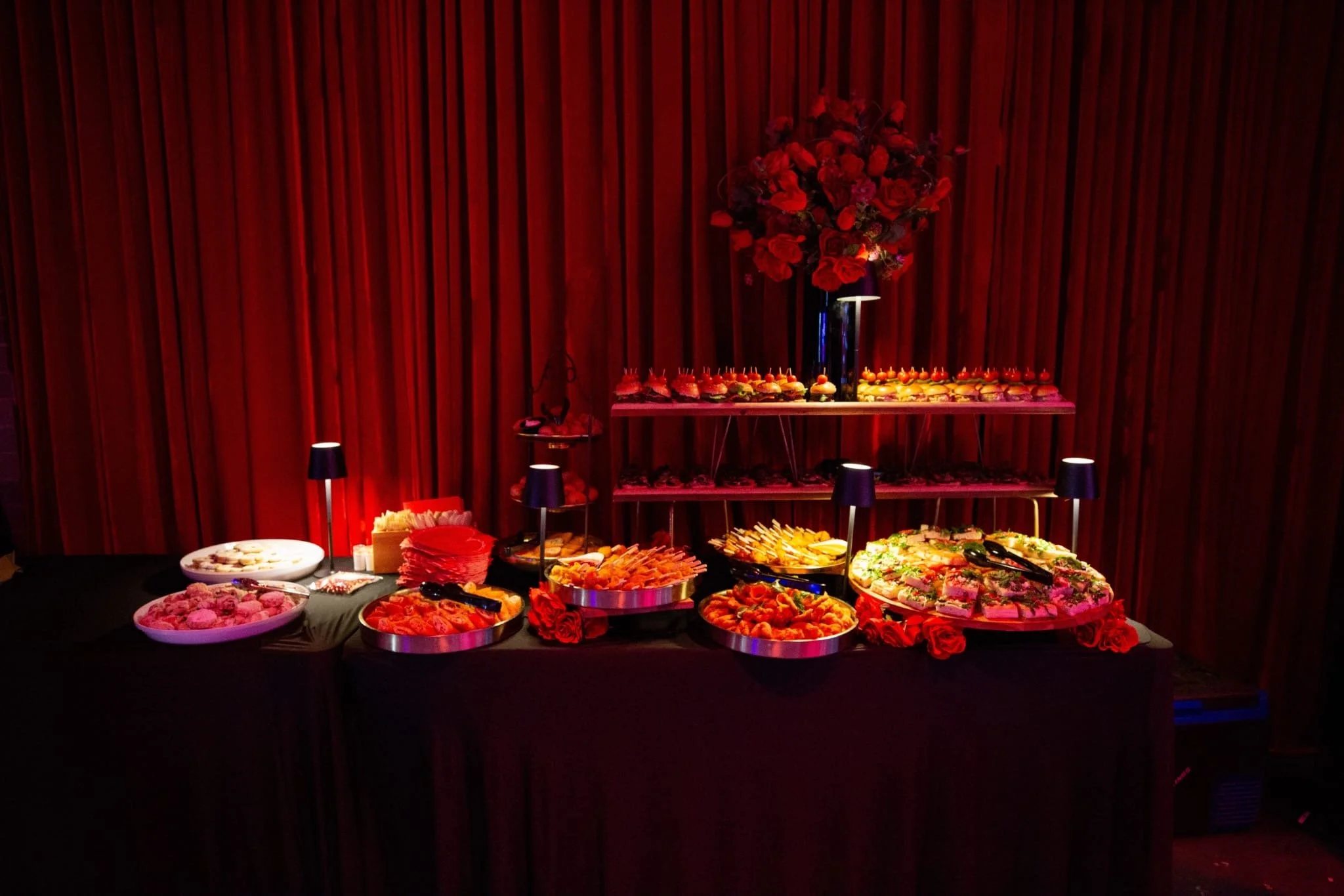A catered buffet table set against a draped red curtain, covered with a black tablecloth. The table is laden with various appetizers, including sandwiches, sliders, pastries, and skewers, illuminated by small black table lamps