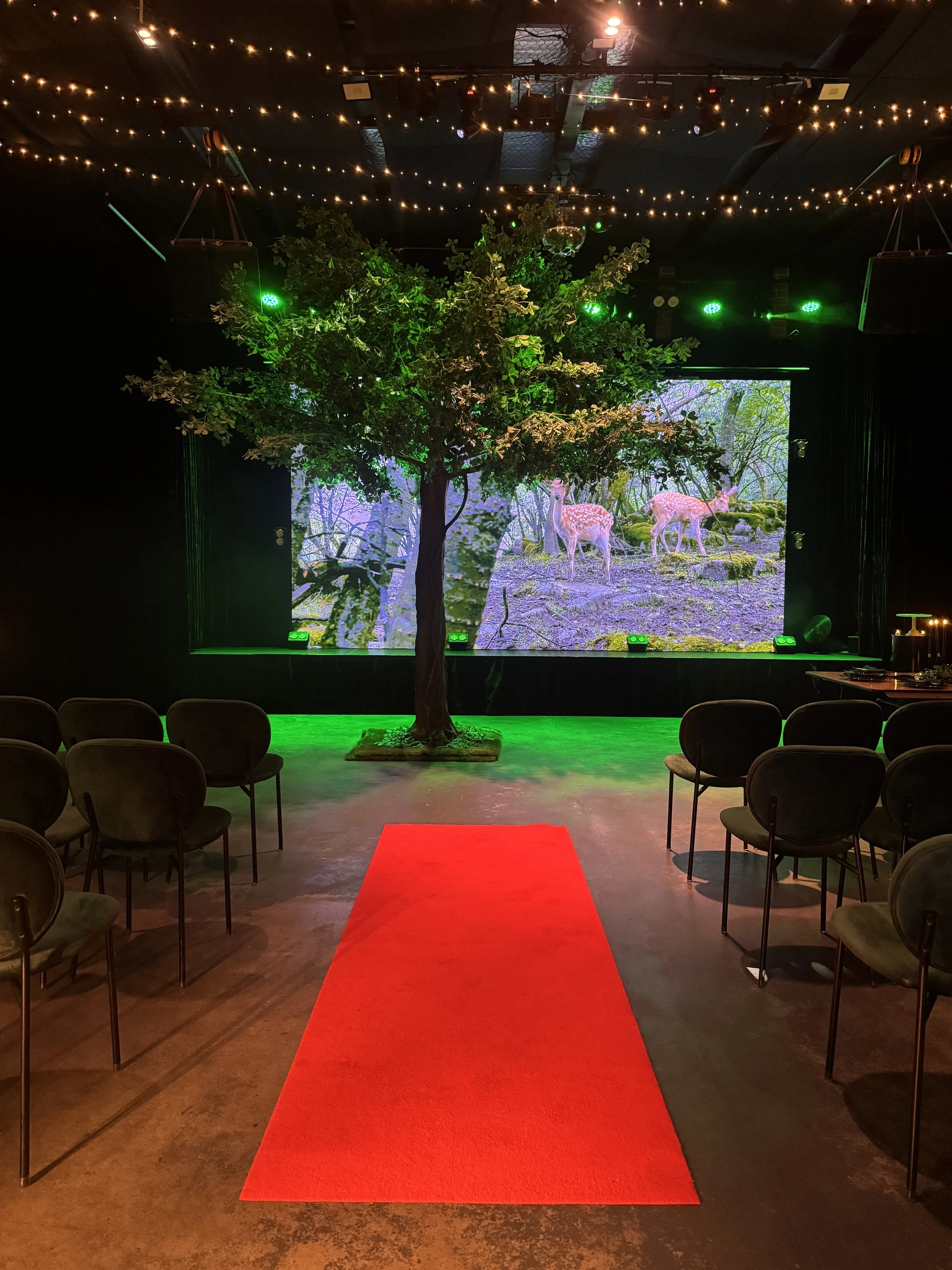 A bright red runner forms a central aisle on the floor, leading toward a stage flanked by an artificial leafy tree. The stage features a digital backdrop of spotted deer in a sunlit forest, with rows of dark green chairs set up on either side.