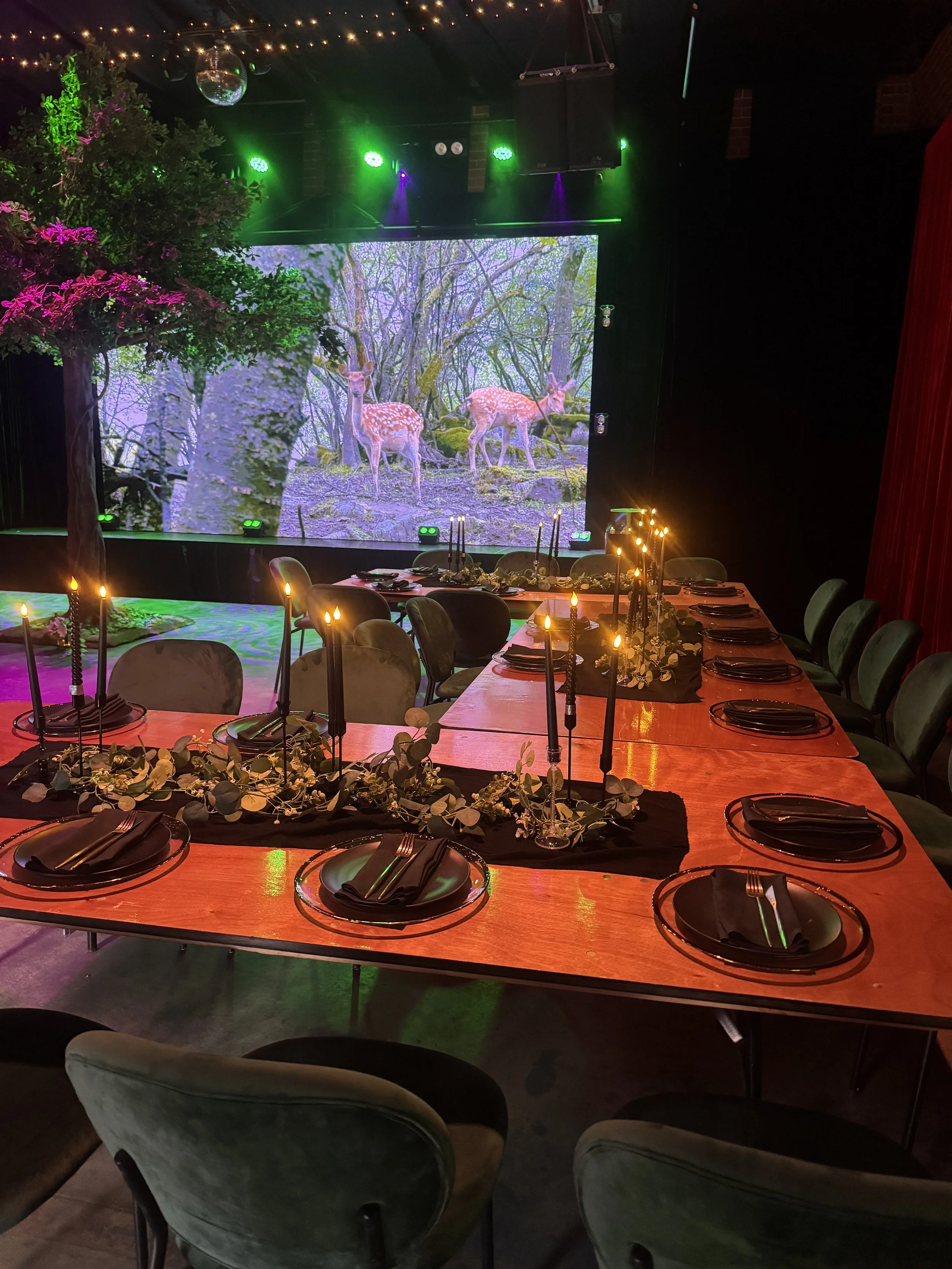 A long wooden banquet table set with black place settings, greenery garlands, and tall black taper candles casting a warm glow. In the background, green lighting highlights an indoor tree prop set against an LED screen showing a woodland forest scene